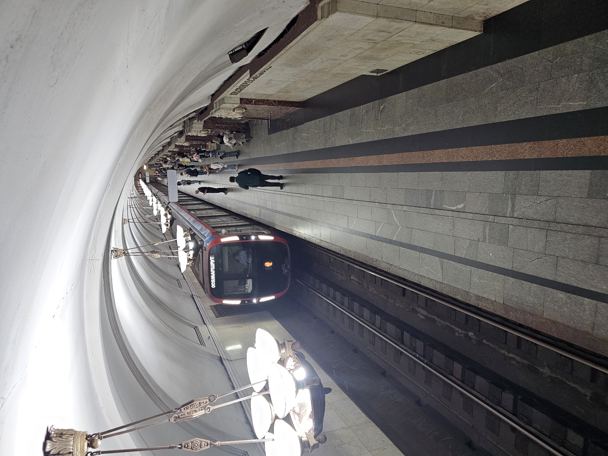 The image depicts the interior of the Krasnopresnenskaya metro station in Moscow, Russia. A red and blue metro train, displaying the Cyrillic text "ООО МАРШРУТ", is stopped at the platform. Several passengers are visible on the platform, some standing and waiting, while others are walking. A man in a dark green jacket and dark pants is standing on the platform facing away from the camera, looking towards the train. The station features a distinctive arched ceiling and ornate chandeliers. The platform is paved with stone tiles and bordered by a darker strip. The walls of the station are adorned with decorative elements and appear to be made of stone. The overall scene captures a typical moment in a busy Moscow metro station.
