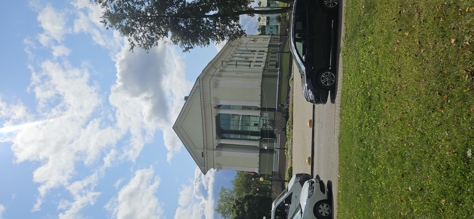 The image shows a pale green neoclassical building with large windows and tall columns, likely a cultural institution, located in Vitebsk, Belarus. The building is situated across from a street with parked cars and a grassy area. A bright sunbeam pierces through a cloudy blue sky, casting light on the scene. The surrounding environment appears to be a public space with trees and a sidewalk.