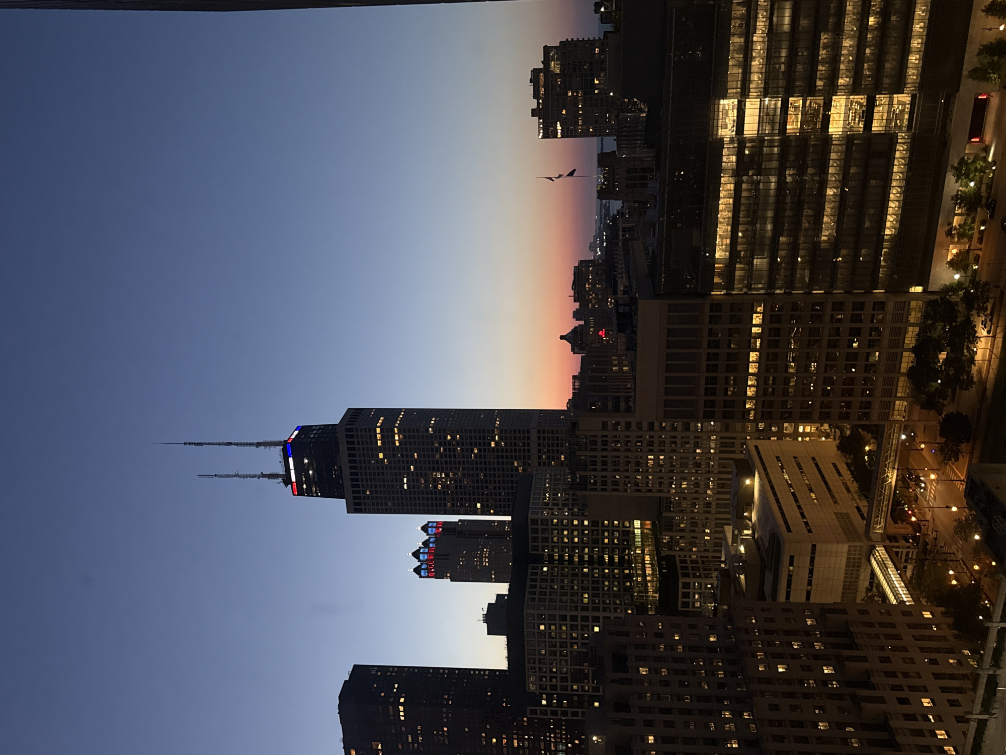 This is a wide-angle, medium-shot photograph of the Chicago skyline at dusk. The sky transitions from a deep indigo at the top to a soft orange and pink hue near the horizon, indicating the sun has recently set. Multiple skyscrapers dominate the scene, with illuminated windows suggesting the buildings are occupied.
The most prominent building is the John Hancock Center, identifiable by its distinctive shape and the red and blue lights near its apex. To the left of the Hancock Center, another tall skyscraper is visible, also lit up with numerous windows. In the middle ground, a cluster of buildings forms a dense urban landscape. One building in this cluster has illuminated red and blue lights forming a pattern on its upper facade. Further in the distance, city lights and the faint outlines of more structures can be seen, hinting at the vastness of the city.
In the foreground, parts of other buildings are visible, appearing darker and less illuminated than those in the mid-ground and background. The overall atmosphere is one of a bustling metropolis transitioning from day to night. The image captures a quintessential view of Chicago's architectural grandeur.