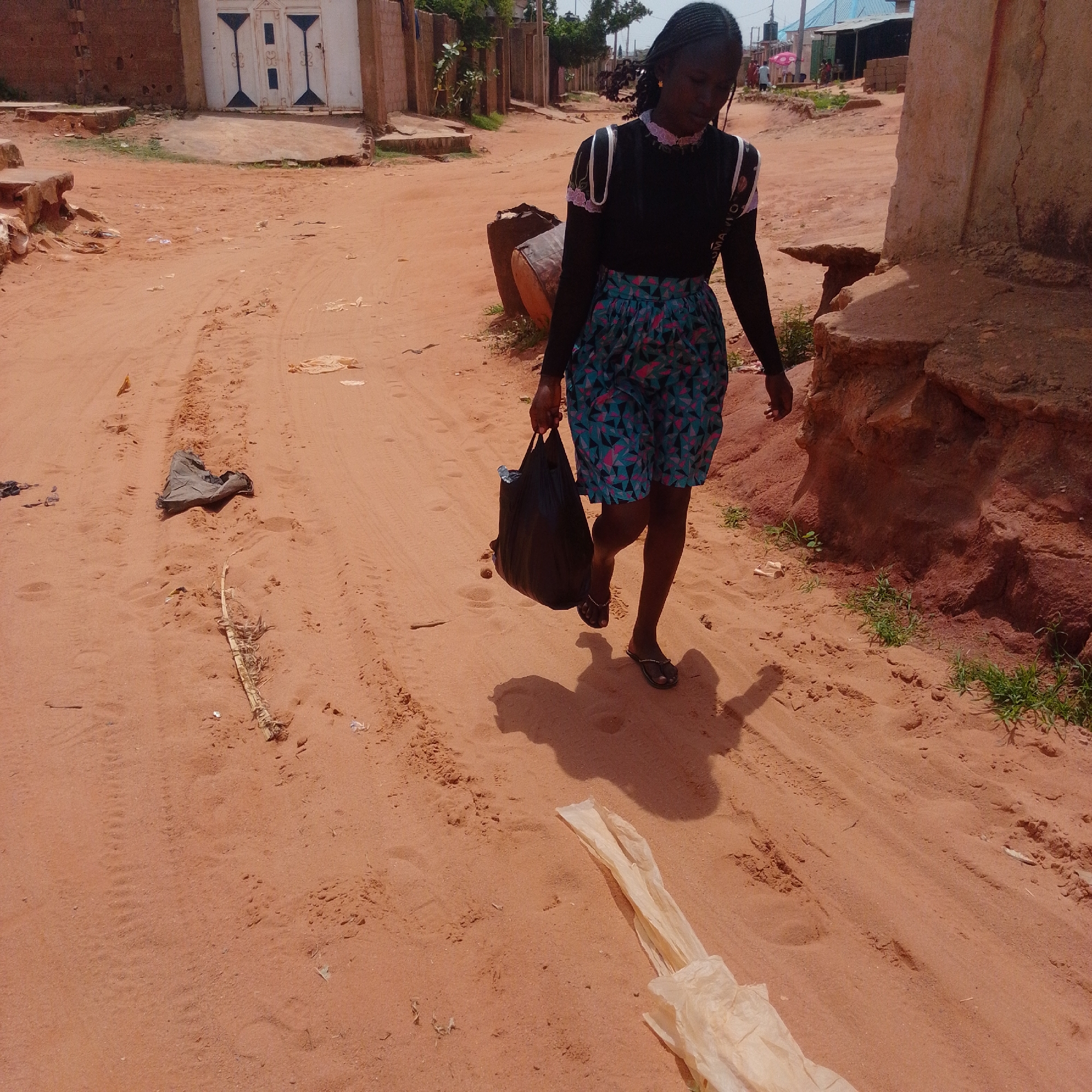 In a dusty, reddish-brown dirt road, a young Black woman walks with a black plastic bag in her right hand. She has dark skin, dark braided hair, and is wearing a black long-sleeved shirt with a decorative pink and white ruffled neckline and cuffs, and a blue, green, and pink geometric-patterned skirt. Her shadow stretches out before her on the sandy ground. The scene appears to be in a rural or semi-urban area, possibly a village in Nigeria given the location context.
In the background, there are simple, low-rise buildings, one with a white facade and ornate dark metal doors. The ground is littered with some debris, including a piece of light-colored plastic or fabric, a dry stick, and some scattered trash. A large, dark cylindrical object, possibly a barrel or drum, rests on the side of the road behind the woman. The lighting suggests it is daytime, with a bright sun casting distinct shadows. The overall atmosphere is one of everyday life in a warm climate.
