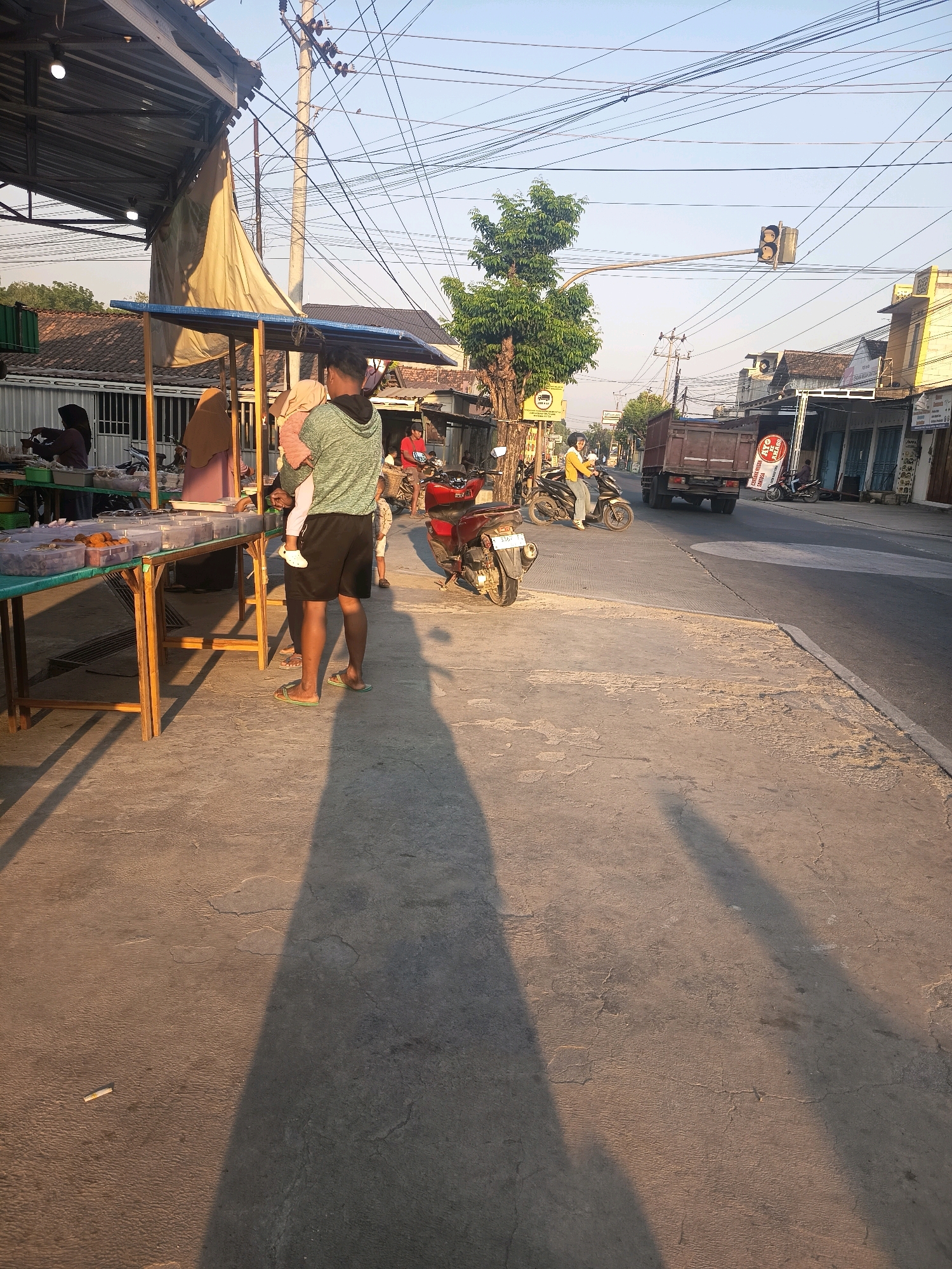 The image depicts an outdoor scene near a roadside market in Pulokulon, Indonesia. In the foreground, a man in a grey hoodie and black shorts stands facing away from the camera, holding a small child. He is casting a long shadow on the concrete ground. To his left, a market stall is visible with a green tarp-covered table displaying food items in clear containers. Several people, including a woman in a hijab, are attending to the stall. In the mid-ground, a red motorcycle is parked on the right side of the road. Further down the street, a truck is driving away, and a person on a motorcycle is approaching from the opposite direction. The background shows buildings lining the street and a clear blue sky with electrical wires crisscrossing overhead. The lighting suggests it is late afternoon or early morning due to the long shadows.