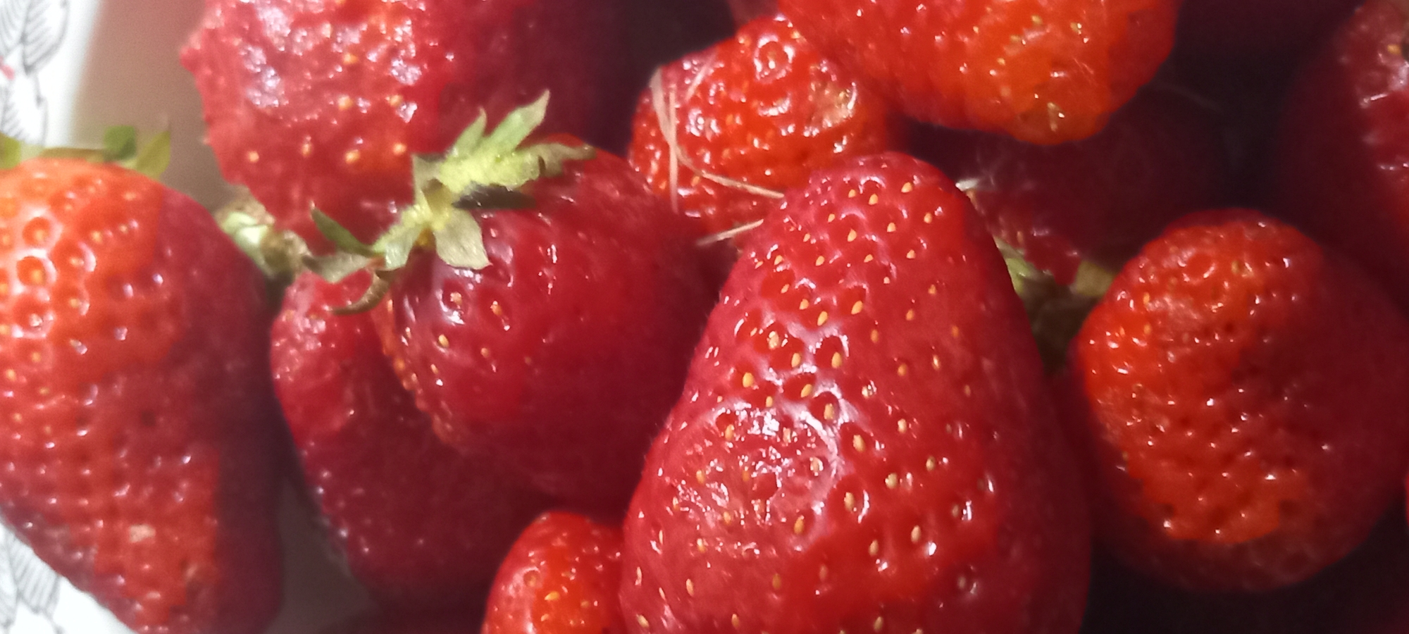 A close-up, overhead shot shows a pile of ripe strawberries. The berries are a vibrant red, with visible seeds dotting their surfaces. Some have green stems attached. The lighting creates highlights and shadows, emphasizing the texture of the fruit. The background is a dark blur, with a hint of a patterned surface at the top left, possibly a bowl or plate. There are no people or recognizable landmarks from Kryvyi Rih, Ukraine, visible in the image.