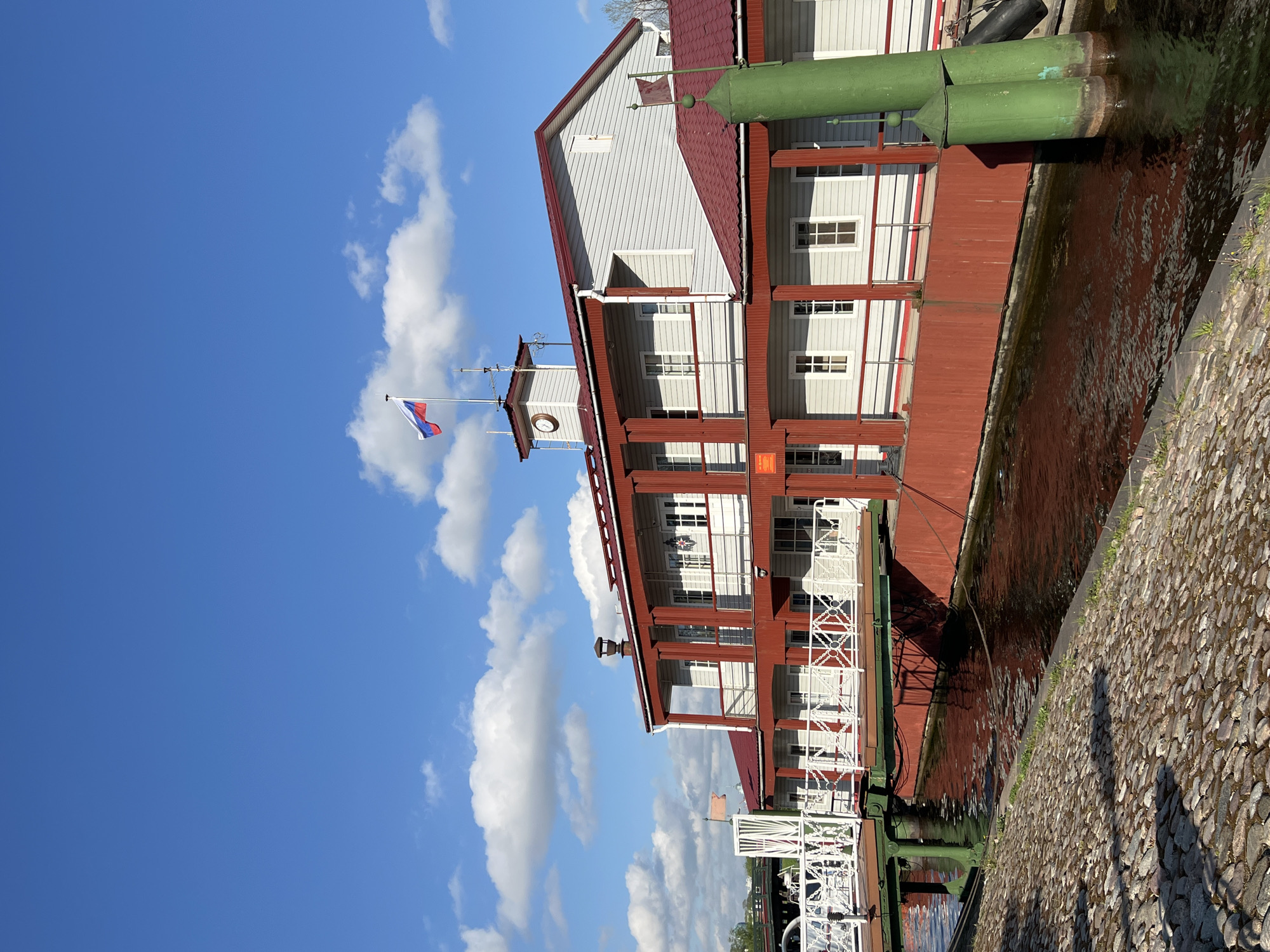 The image shows a three-story building with a red roof and white siding located on the edge of a canal. The building features multiple windows and balconies, and it appears to be constructed in a traditional style. A Russian flag is visible atop a tower on the building. The canal reflects the building and sky. The bank of the canal is lined with cobblestones. The location is likely Saint Petersburg, Russia, given the architectural style and flag.