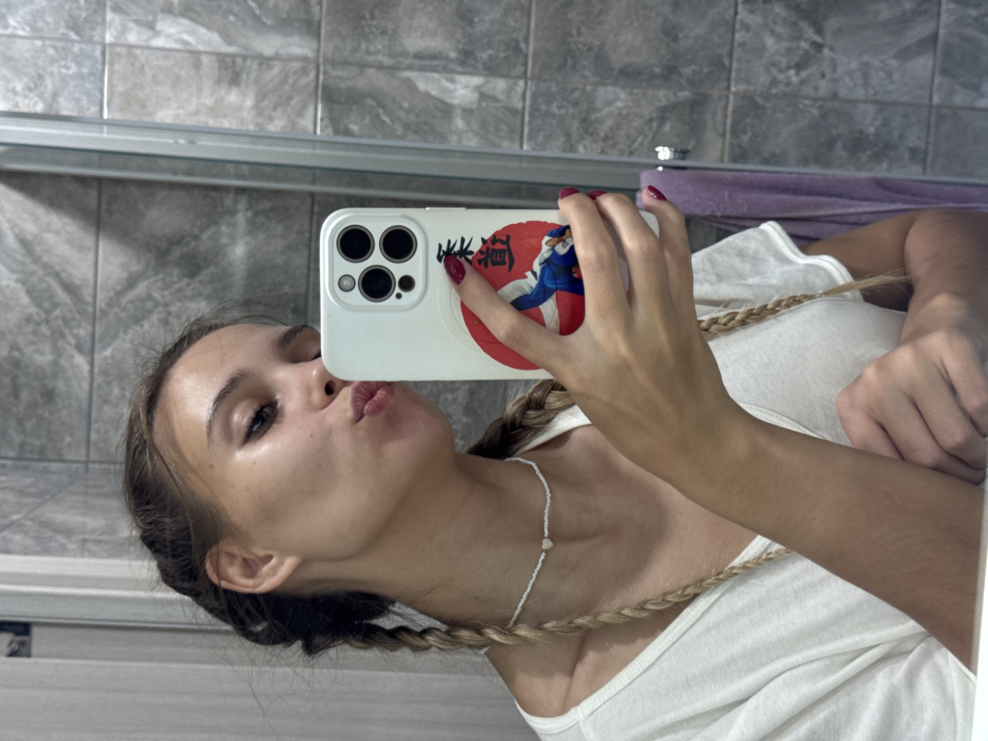 In a bathroom in Chaikovsky, Russia, a young adult female subject captures a mirror selfie. She has light brown hair styled in two braids, wears a white V-neck t-shirt, and a white beaded choker necklace with a central heart-shaped bead. Her lips are puckered in a pout. She holds a white smartphone with a visible three-lens camera module. The phone case features a red circular graphic depicting a person in a blue martial arts gi, with two Japanese characters (柔道 - Jūdō) printed vertically above it. Her fingernails are painted dark red. The background, reflected in the mirror, consists of grey-toned tiled walls, a partial view of a glass shower enclosure, and a purple towel hanging on a rack.