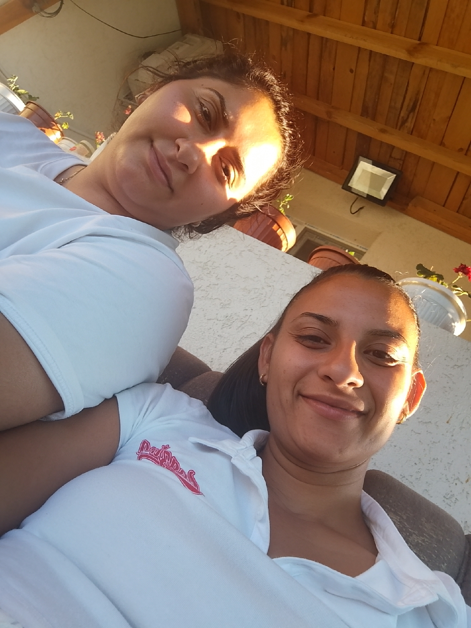 Two individuals are captured in a selfie, positioned in a close-up shot outdoors. The person in the foreground, on the right, is a woman with dark hair pulled back and wearing a white polo shirt with a red logo. She is smiling, with her head tilted and looking towards the camera. The second individual, in the background on the left, is partially visible, also appearing to be a woman with dark hair, wearing a white shirt and a necklace. She is looking slightly away from the camera, with sunlight illuminating her face.
The environment suggests an outdoor setting with a textured white wall in the background. Above the wall, a wooden ceiling structure is visible, along with potted plants, some containing red flowers. A black light fixture is mounted on the wooden ceiling. The lighting indicates it is daytime, with the sun casting a warm glow. The overall scene conveys a casual and relaxed atmosphere. It is stated that this scene is located in the city of Zimnitsa, Bulgaria.