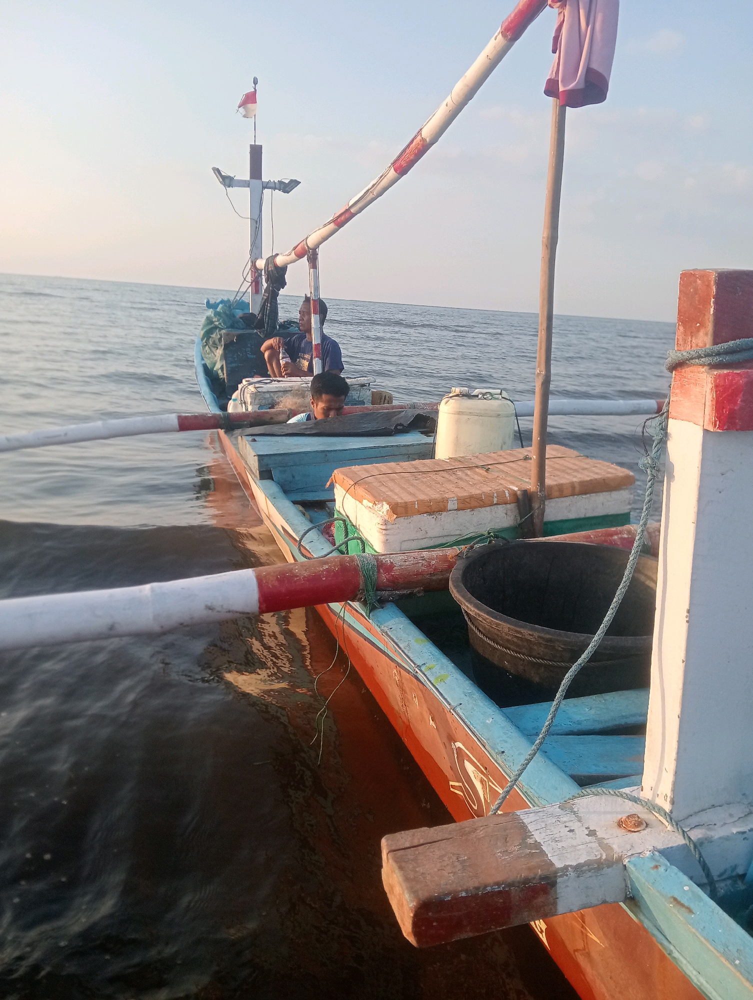 The image captures a close-up view of a wooden fishing boat at sea, likely in Pesisir, Indonesia, based on the context provided. Two individuals are visible on the boat. One man, wearing a dark blue shirt, is seated and looking towards the right. Another person, partially obscured and looking downwards, is seated further into the boat. The boat is equipped with fishing gear, including a large black basket, several storage containers, and what appear to be oars or poles extending from the sides. A Indonesian flag is planted on a mast at the front of the boat. The sea is calm, with a clear sky suggesting either early morning or late afternoon light. The boat itself exhibits signs of wear, with peeling paint in places and weathered wood. The overall scene depicts a moment of stillness on a working fishing vessel.