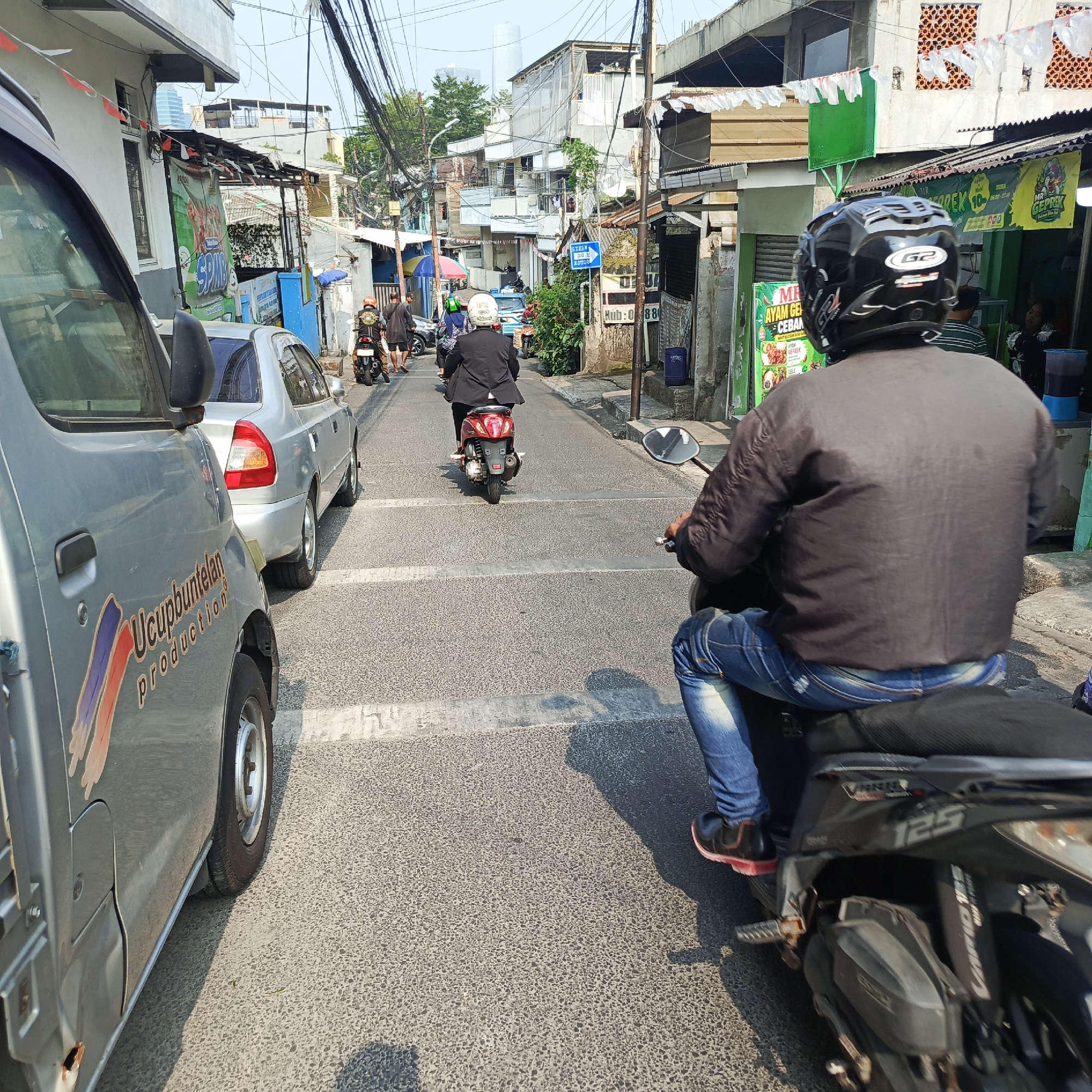 The image depicts a street scene in South Jakarta, Indonesia, on a bright, sunny day. The perspective is from behind a silver van on the left side of the frame, looking down a narrow, asphalt street lined with buildings. Several motorcycles and a silver sedan are present, suggesting traffic.
In the foreground, a person on a black motorcycle wearing a black helmet with a "G2" logo and blue jeans is visible, facing away from the camera. The motorcycle has "125" visible on its side. To the left of this motorcycle, the side of the silver van is prominent, showing a logo with the text "Ucupbuntelan production".
Further down the street, another motorcycle is seen with a rider in a black jacket and white helmet, and a red scooter is also visible with a rider. On the right side of the street, there are various buildings, some appearing to be shops or food stalls with banners, including one for "MR. GEPREK". People are walking on the sidewalk on the right. Power lines crisscross overhead, and some laundry is hanging from buildings. The atmosphere is that of a bustling, ordinary urban environment.