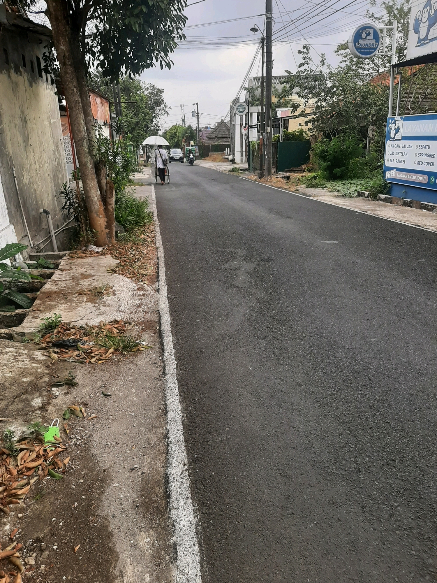 A person pushes a bicycle with an umbrella in the city of Sumampir, Indonesia. The scene appears to be a residential street with a sidewalk, a road, and buildings on either side. The sky is overcast. A laundry service sign is visible on the right side of the street. The foreground shows a sidewalk with fallen leaves and a green plastic bag.