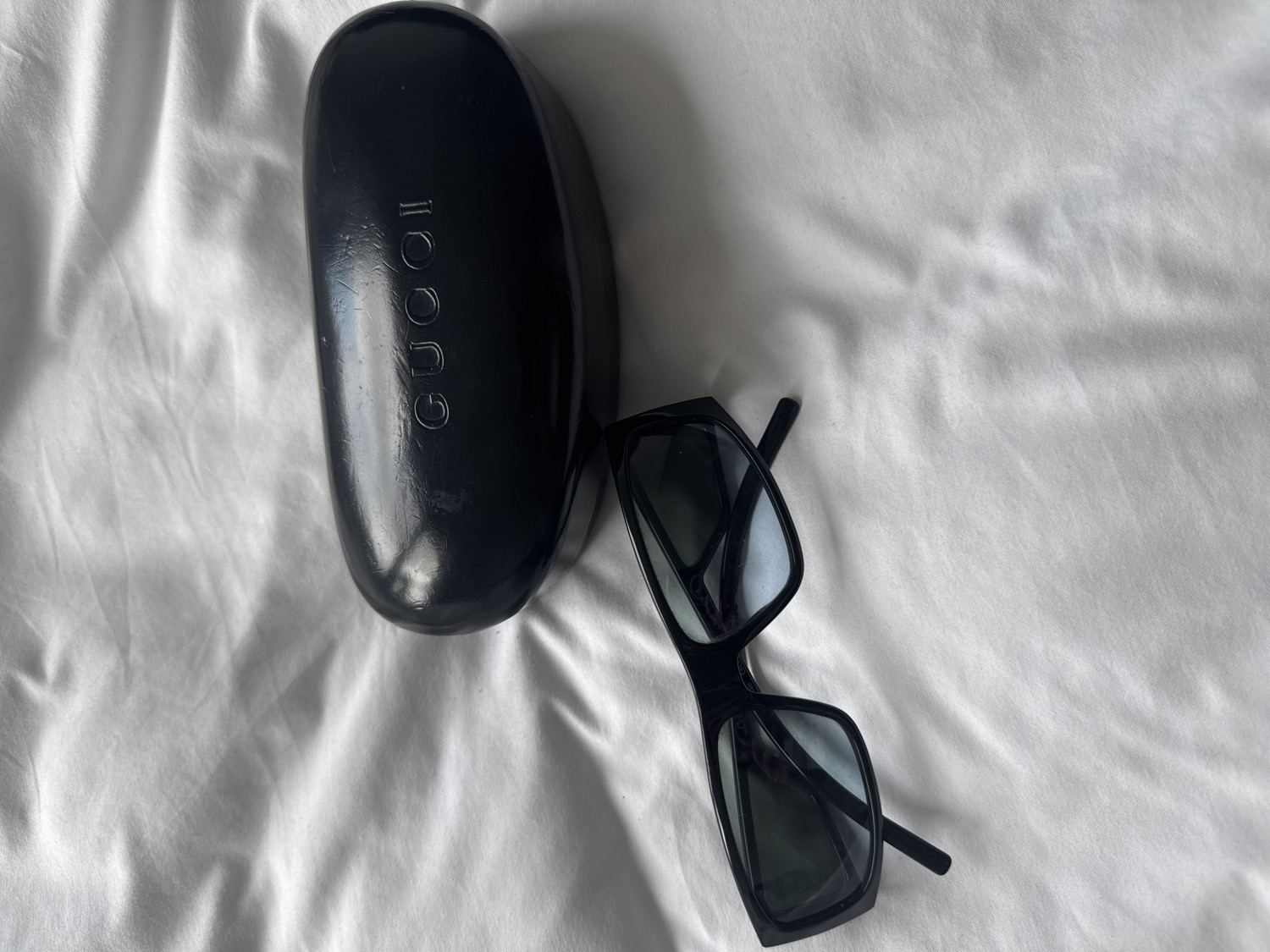 This overhead shot displays a black sunglasses case and a pair of black sunglasses resting on a white, wrinkled fabric surface, likely a bedsheet or tablecloth. The setting is indoors, with soft, diffuse lighting that suggests either natural light or ambient room illumination, casting subtle shadows.
The primary subjects are the two objects. The sunglasses case is a glossy black, oval-shaped hard case. The brand name "GUCCI" is prominently embossed or debossed vertically on its top surface. The case appears to have some minor wear or dust on its reflective surface. To the right of the case, the black-framed sunglasses are positioned at an angle, with their arms extended. The frames are rectangular or slightly square, and the lenses are dark, reflecting some light from the environment.
There is no specific activity or event taking place; the image is a still life composition of these items. No people are visible, and no emotions are conveyed. The location context provided is London, United Kingdom, although there are no visual cues within the image itself to confirm this. The text "GUCCI" is the only visible writing in the frame, clearly identifying the brand of the items.