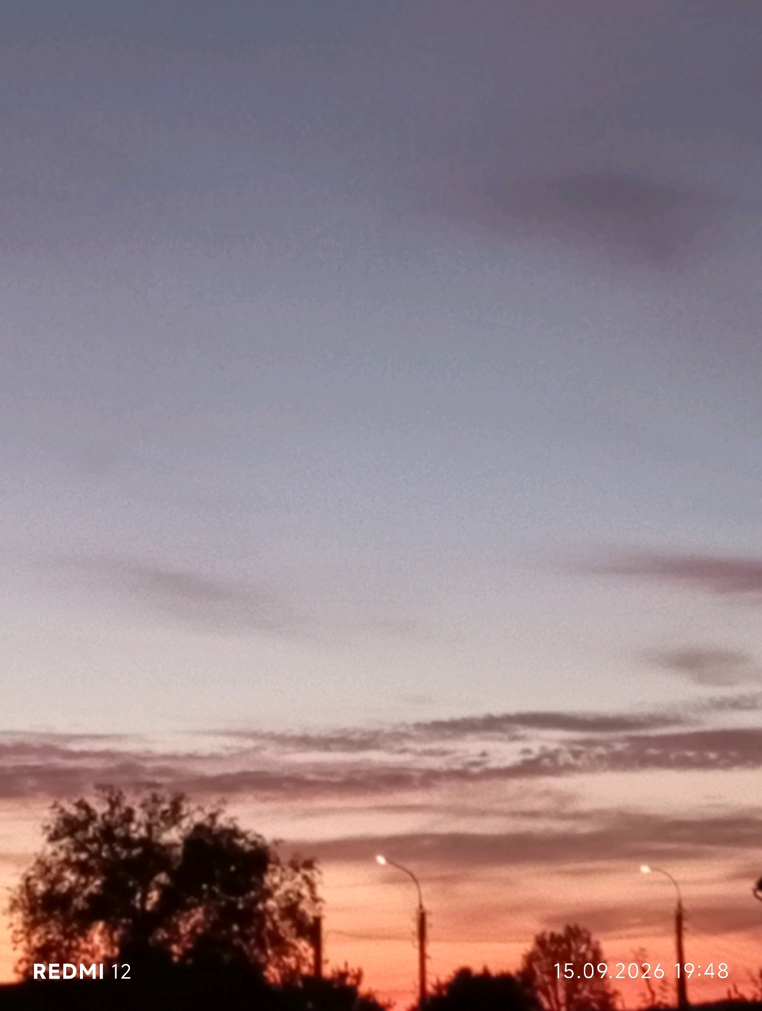 The image shows a sunset scene with a dusky sky in Smila, Ukraine. In the foreground, silhouetted trees and streetlights are visible against the vibrant orange and pink hues of the sunset. The date stamp "15.09.2026 19:48" and "REDMI 12" are present in the lower corners of the image. The overall atmosphere is calm and serene.