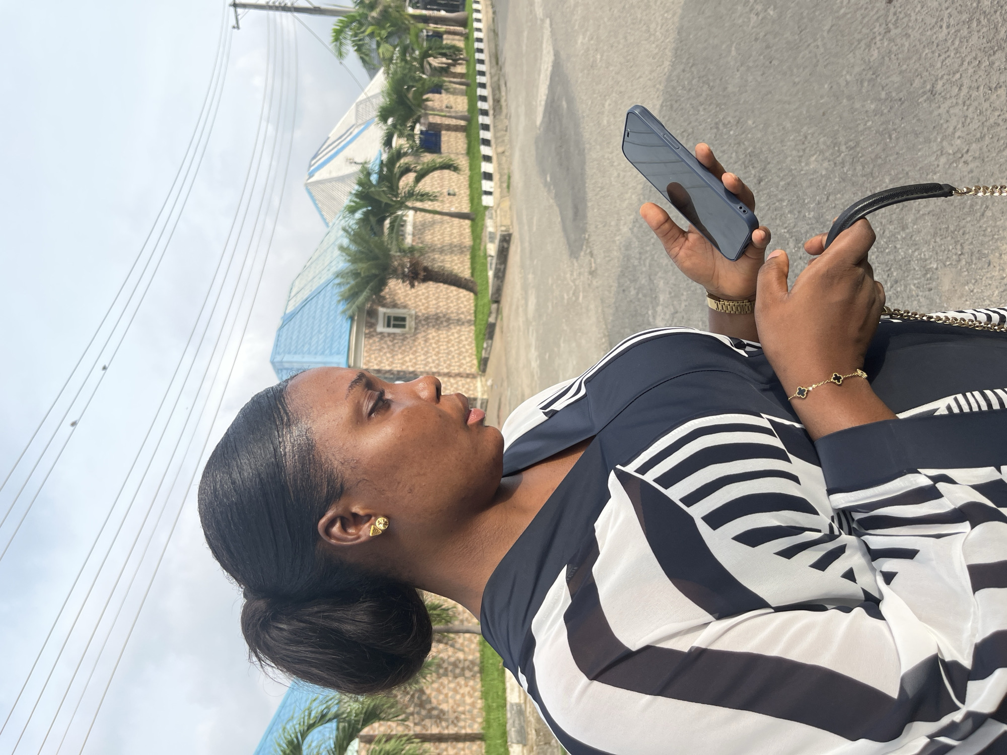 A person is looking at a blue phone in their hands on a paved road in Abuja, Nigeria. The person is wearing a black and white striped top, a gold watch, and a gold bracelet, and they are holding a black purse with a gold chain. Palm trees line the road behind the person, and a low building with a blue roof can be seen in the background. Overhead power lines are visible against a cloudy sky.