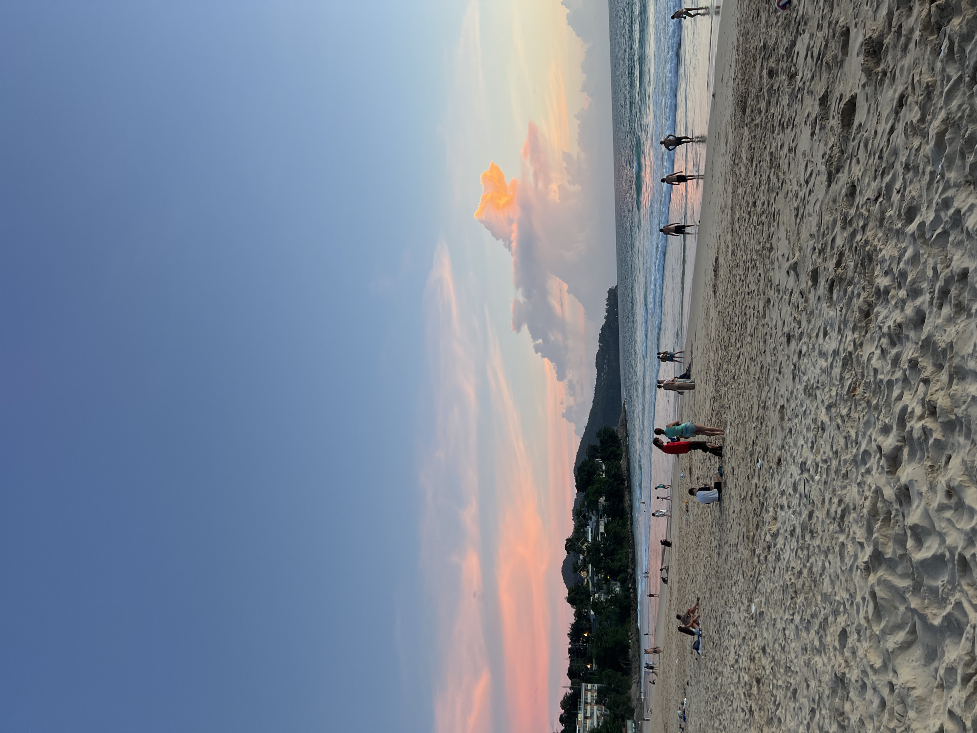 The image depicts a beach scene at sunset in Choeng Thale, Thailand. The sky is a gradient of blue and pink, with fluffy clouds illuminated by the setting sun. In the background, a lush green coastline is dotted with buildings. The beach itself is sandy, with gentle waves lapping at the shore. Several people are visible on the beach and in the shallow water. Some are walking along the shoreline, while others are gathered in small groups. A few individuals appear to be playing in the water. The overall atmosphere is serene and relaxed, typical of a popular beach destination.
