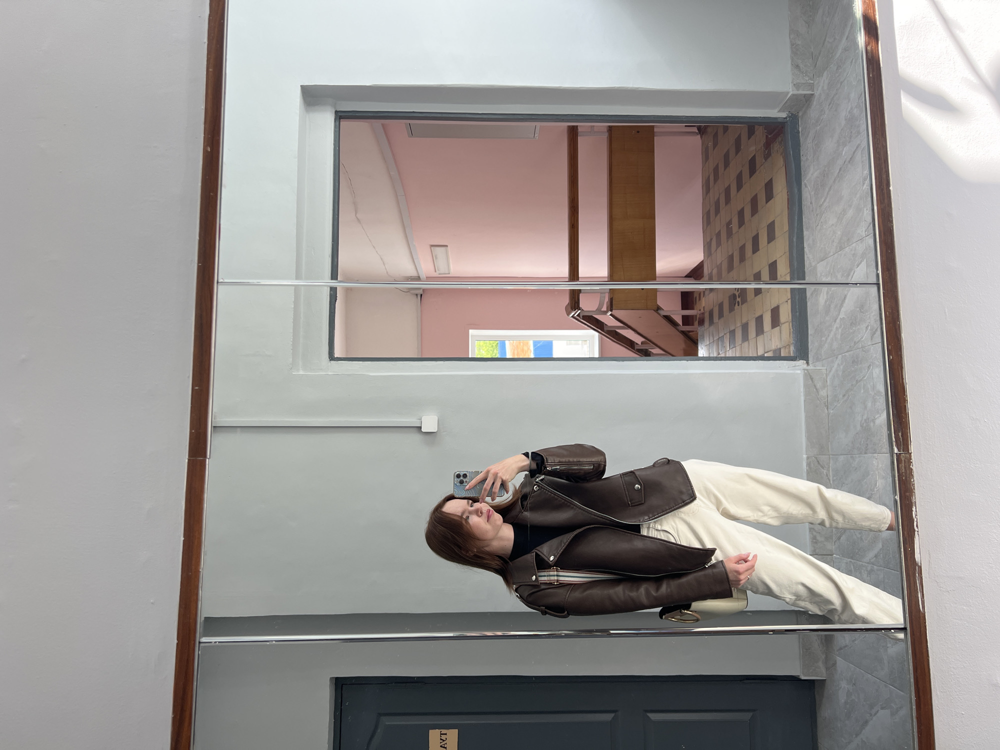 This media file captures a mirror selfie taken indoors in Chaikovsky, Russia, with the image rotated 90 degrees to the right. A young woman with shoulder-length brown hair is reflected in a full-length mirror, appearing to lie horizontally as she holds up a dark-colored iPhone (likely a Pro model) to take the picture.
She is dressed in a dark brown leather moto jacket over a black top and cream or off-white wide-leg pants. A small white shoulder bag is worn across her body. Her left hand is visible near the bottom of her jacket, holding the strap of her bag.
The mirror reflects a multi-layered interior. Above the woman, a large rectangular opening reveals a room with light pink walls and a white ceiling, featuring a rectangular light fixture and another smaller light. Further right in this reflection, a wooden column and what appears to be part of a metal staircase are visible. Below this section, a window in the reflected room shows glimpses of green foliage outside.
To the right of the woman in the reflection, a wall with a distinctive brown and off-white checkered tile pattern is visible. A gray tiled wall borders the right side of the mirror itself, while a plain light gray wall runs along the left. A horizontal silver bar spans across the mirror's width, roughly at the woman's waist level. At the very bottom of the mirror, part of a dark door is reflected, displaying the Cyrillic text "АУТ" (meaning "OUT") on a light-colored sign. The mirror is framed by thin dark wooden borders.