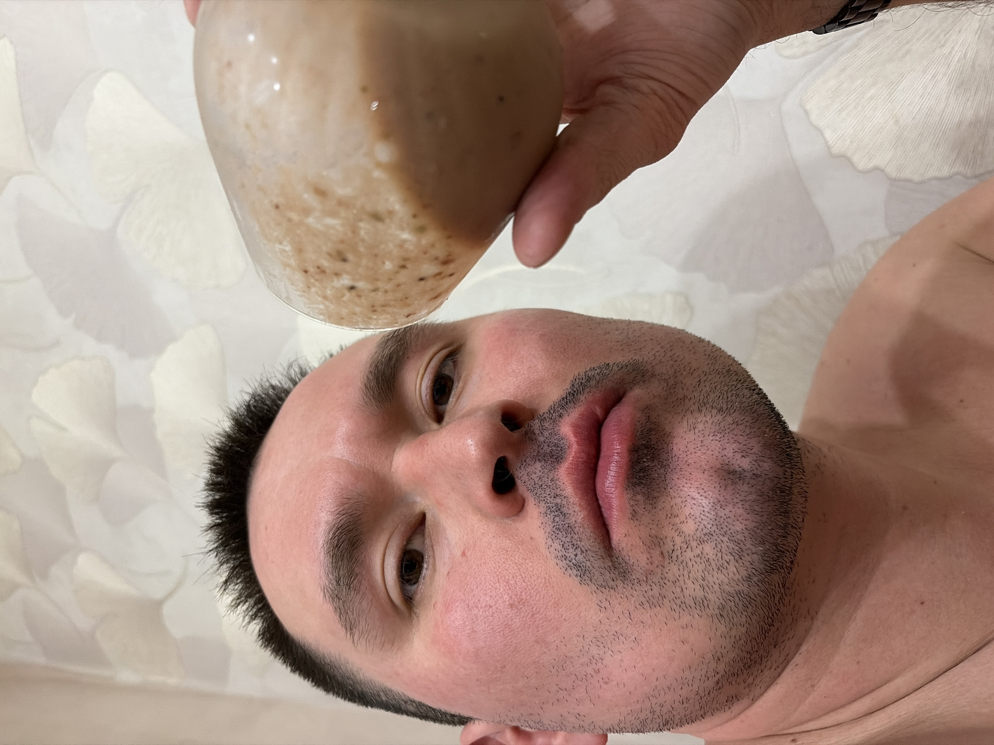 A man is holding a glass of liquid above his head. The liquid appears to be a smoothie or shake. He is looking up towards the glass with his mouth slightly open. His facial hair is dark and his complexion is fair. He appears to be shirtless. The background is a light-colored, patterned wall. It is unlikely to determine a specific city or location from these details.