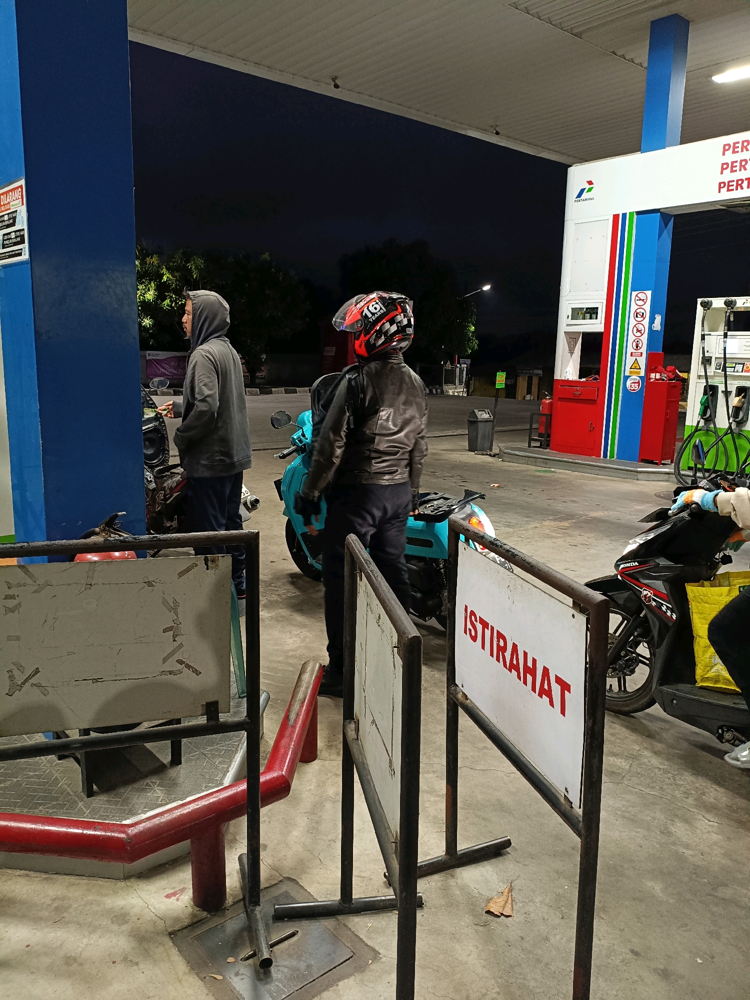 The image depicts a night scene at a Pertamina gas station in Maguwoharjo, Indonesia. Two individuals are present. One person, wearing a grey hoodie and dark pants, stands near a blue pillar, seemingly interacting with a motorcycle. Another individual, wearing a black leather jacket and black pants, with a red and black helmet, stands beside a turquoise scooter, facing away from the camera. In the background, Pertamina signage is visible on the gas station structure. Several motorcycles are parked in the vicinity. The ground is concrete, and the sky is dark. A white sign with red text reading "ISTIRAHAT" (Indonesian for "REST") is prominently displayed in the foreground.