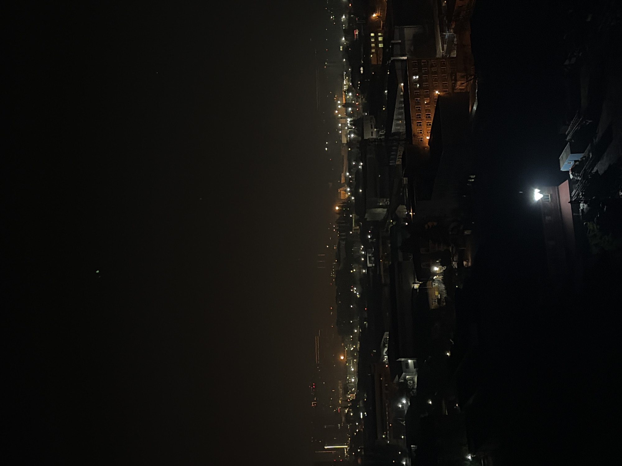 The image, when viewed in its intended landscape orientation (rotated 90 degrees clockwise from the provided original), captures a night-time cityscape or townscape from an elevated perspective.
The scene is divided horizontally by a prominent line of artificial lights. The upper portion of the image features a dark, hazy night sky, with one small, distinct greenish light dot visible. The middle ground is dominated by a sprawling array of warm-toned streetlights and building lights, stretching across the horizon. This band of lights suggests a street network or the illuminated edge of a city along a significant feature.
The lower right section of the image (when rotated) is a very dark expanse, possibly a body of water such as a river or bay, reflecting some distant light sources very faintly. To the lower left, a denser cluster of buildings is visible, with numerous illuminated windows casting a warm glow. One large building with multiple rows of glowing windows stands out. The artificial lighting, primarily yellow and orange, creates a vibrant yet hazy atmosphere, characteristic of light pollution in an urban environment at night.
The time of day is clearly night. No specific activities or people are discernible. The overall impression is of a sprawling city at rest, illuminated by its own infrastructure. There is no visible text in the image. The location context provided is Krasnodar, Russia.
