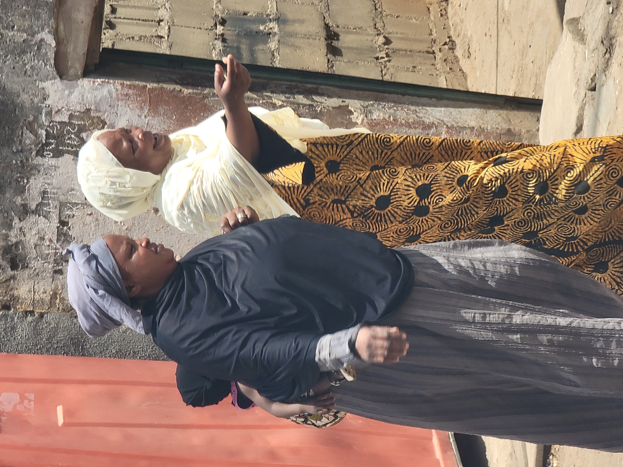 Two women are captured in what appears to be an outdoor setting in Maiburiji, Nigeria, under bright, clear sunlight, indicating daytime. They are standing side-by-side, suggesting they are together or passing each other.
The woman on the right (facing the viewer's left if the image were upright) wears a light yellow head covering (possibly a hijab or scarf) and a matching light yellow, pleated top or dress. Beneath this, a dark top is visible. Her skirt is a distinctive dark yellow or gold with a repeating black pattern resembling abstract suns or spirals. She is looking upwards with a joyful or amused expression, her mouth open in a smile or laugh, and her right arm is slightly raised. She wears a ring on her left hand.
The woman on the left (facing the viewer's right) wears a light gray or blue head covering and a black long-sleeved top or dress. Her skirt is dark gray, possibly textured or pleated. Her expression is more neutral or contemplative, and her hands are clasped in front of her or holding something out of view.
The background features a rough, textured wall, possibly made of concrete or brick, showing signs of wear. In the foreground, a reddish-orange surface, possibly a metal container or part of a building, occupies the lower portion of the image. There is no visible text. The women's attire is modest and consistent with clothing styles often seen in the region.