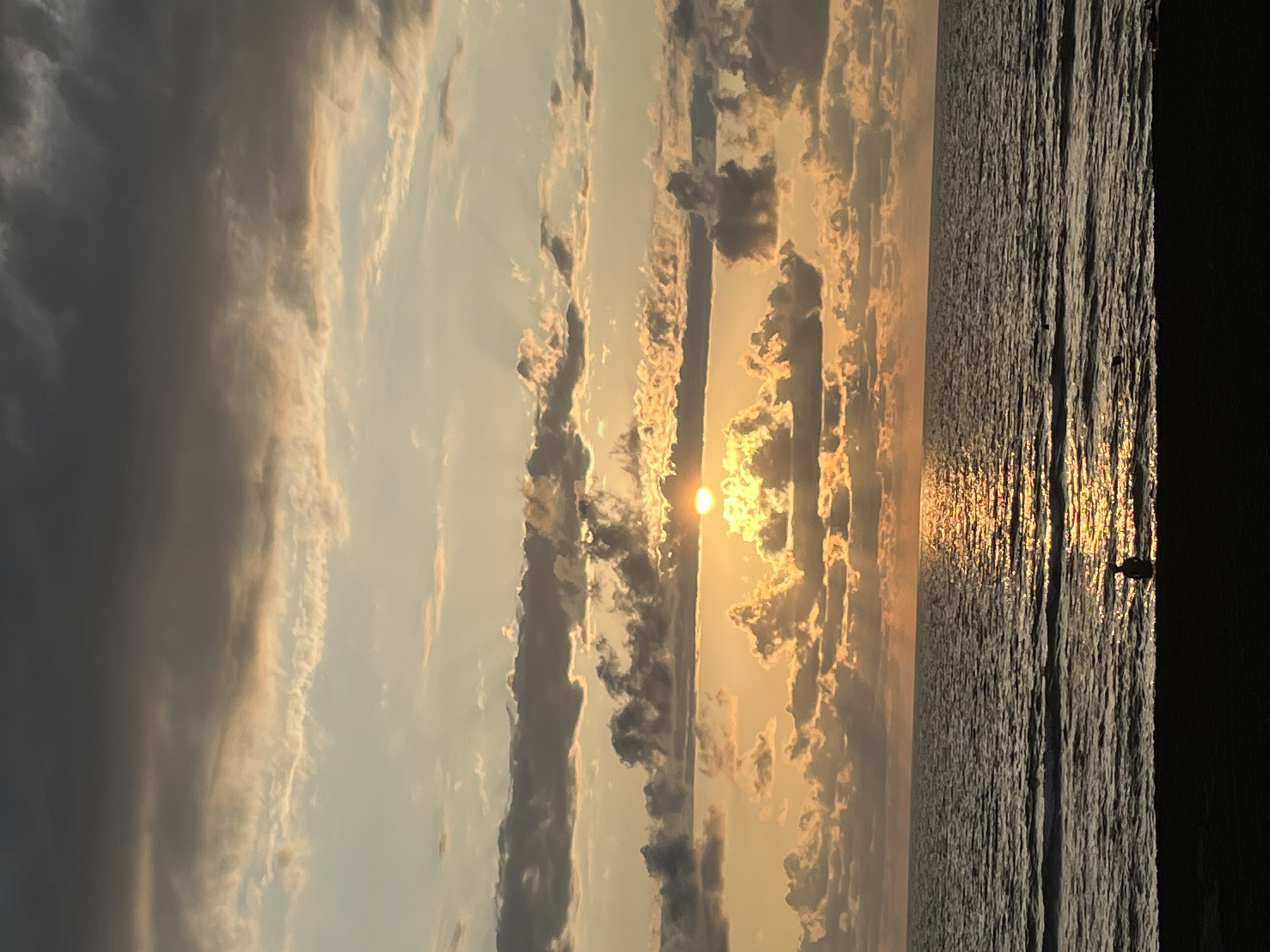 This photograph captures a sunset over the Black Sea coast in Ureki, Georgia. The sky is filled with a mix of dark gray and lighter, orange-tinged clouds, with the sun partially obscured by a layer of clouds, casting a warm glow and its reflection on the water. The sea is calm with gentle waves. In the foreground, a dark silhouette of a person is visible sitting near the water's edge on a sandy or dark beach. The overall scene conveys a sense of tranquility.
