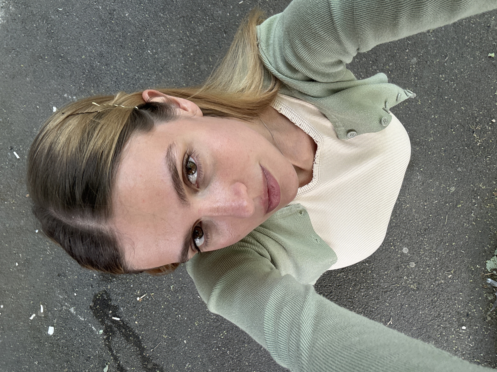 A selfie-style photograph taken from a low angle depicts a young woman standing on a gray asphalt surface, presumably a sidewalk or street. The woman has light brown hair with darker roots, styled with several bobby pins visible near her left ear. She is wearing a light green cardigan over a light beige ribbed top. The background consists of the asphalt surface, which shows scattered debris, including what appears to be small pieces of paper or wrappers. The location is likely outdoors in a city setting. The exact location within Tbilisi, Georgia is not discernible from the image.