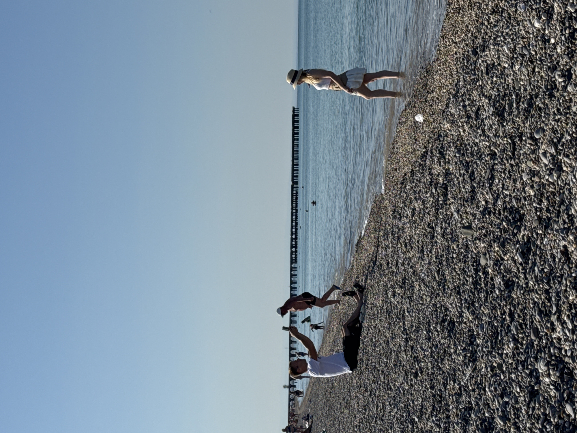 The image depicts a pebble beach in Novofedorovka, Ukraine. A man in a white t-shirt and black shorts is seated on the pebbles, holding a smartphone and appearing to take a selfie or record a video. In the background, a pier extends into the calm sea. A woman in a white top and a light-colored skirt stands in the shallow water near the shore, looking out towards the sea. Other beachgoers are visible further along the coastline and in the water, with a dark pier structure dominating the horizon. The sky is clear and blue.