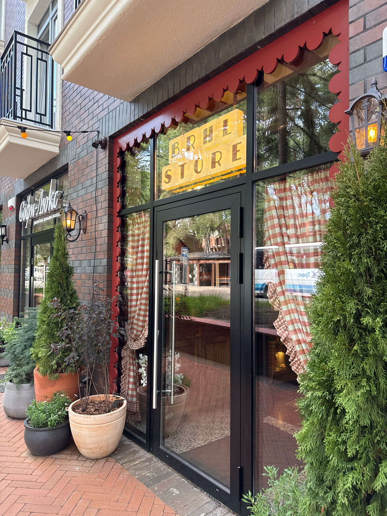 The image shows the exterior of a shop called "BRIHL STORE." The shop has a large glass door and window, framed by a dark material. Above the window, a decorative scalloped red awning is visible. Inside, through the window, red and white checkered curtains are hanging. A yellow sign with dark lettering is prominently displayed in the window, spelling out "BRIHL STORE."
To the left of the main entrance, another window displays the shop's name in white script, "Софи-Люкс" (Sofi-Lyuks), with smaller text below that appears to be "стильные модные аксессуары" (stylish fashion accessories). A vintage-style lantern with a glowing bulb is mounted on the brick wall next to this window.
The building is constructed with brickwork, featuring a mix of grey and reddish-brown bricks. In front of the shop, several potted plants and trees are arranged on a red brick pavement laid in a herringbone pattern. These include a tall, slender evergreen tree to the right of the entrance, a smaller dark-leafed shrub, and other greenery in various pots.
The scene appears to be set during the daytime, with soft natural light suggesting it might be late afternoon or early evening. The reflections in the shop window show trees and some buildings, indicating an outdoor location, likely a street or pedestrian area. The overall impression is that of a quaint, perhaps boutique, store.