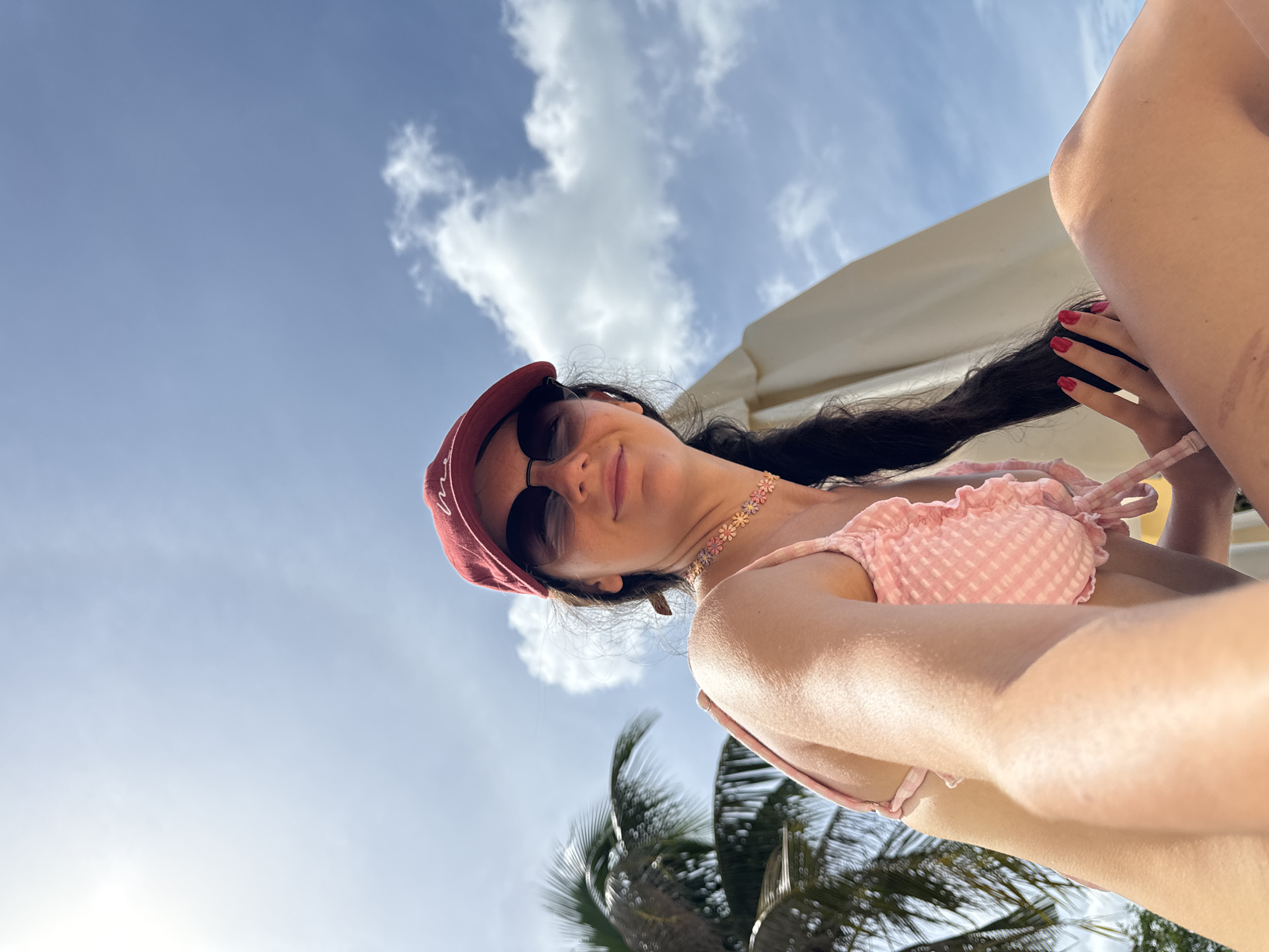 A low-angle, close-up selfie captures a young woman with long, dark hair, wearing a red baseball cap, sunglasses, a pink checkered bikini, and a floral choker necklace. She is seated outdoors with a bright, partly cloudy sky and palm trees visible in the background. A large, light-colored beach umbrella is partially visible behind her. The scene is set on a sunny day, likely at a beach or resort location in Phu Thuy Ward, Vietnam.