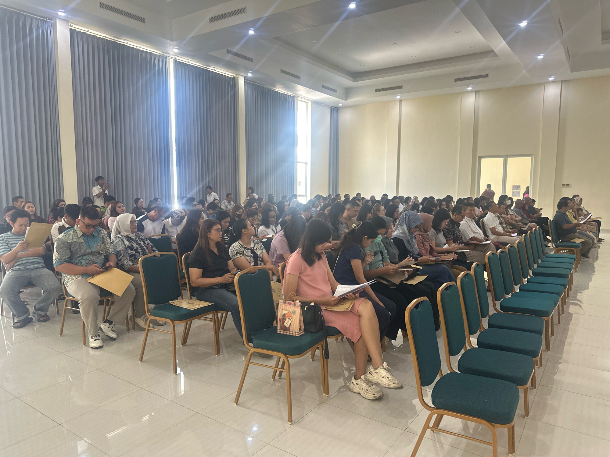 The image depicts a scene inside a large, well-lit hall, possibly a convention center or a lecture hall, in Mojokerto, Indonesia. The hall features a high, coffered ceiling with multiple recessed lights and ventilation grilles. Tall, gray curtains line the left side of the room, extending from ceiling to floor and creating a sense of formality. Natural light streams in from a large window or opening behind the curtains, casting bright rays into the room.
The majority of the image is occupied by rows of chairs arranged in an audience seating configuration. The chairs are upholstered in a dark teal fabric with wooden frames and legs. The floor is a light-colored, reflective tile.
A diverse group of people, predominantly adults, are seated in the chairs, facing towards the unseen front of the hall. Many are looking forward, suggesting they are attending a presentation, lecture, or event. Some individuals are holding papers or books, and a few are engaged in conversation. The attire of the attendees ranges from casual to semi-formal, with some wearing collared shirts and others more relaxed clothing. Several women are wearing headscarves, indicating a likely Muslim majority in attendance.
In the foreground, several rows of empty chairs are visible, indicating the seating arrangement and the potential for more attendees. The lighting is bright and even throughout the hall, with additional accent lighting along the walls. The overall atmosphere appears to be one of organized attendance at a public event.