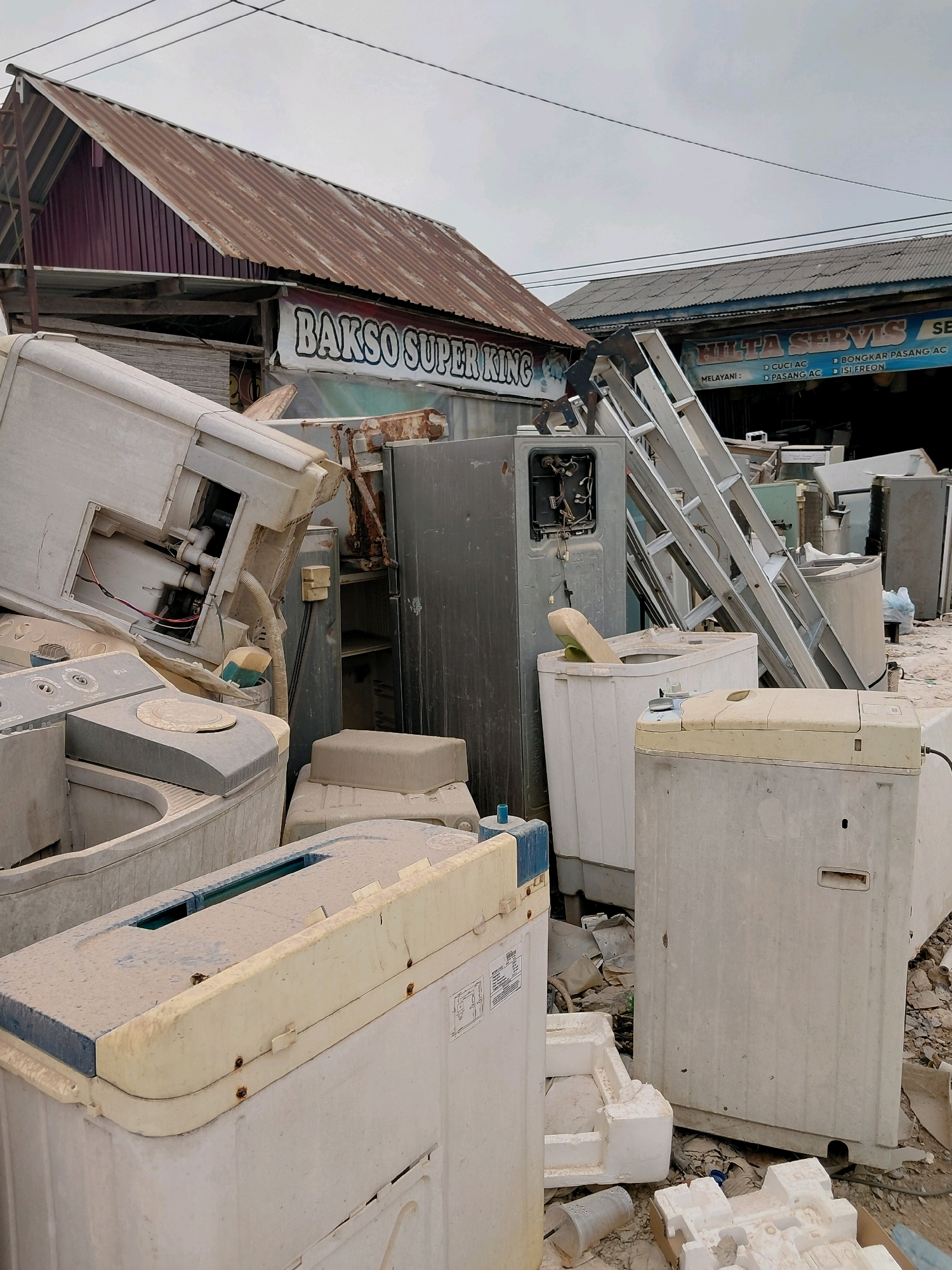 The image shows a junkyard or scrap area, likely in Persiapan Mulya Raya, Indonesia, filled with discarded appliances, primarily washing machines and refrigerators. In the background, a building with a corrugated metal roof, displaying a banner that reads "BAKSO SUPER KING," suggests a commercial area. Another building with a sign for "HILTA SERVIS SE" is also visible, indicating a service-related business. A silver aluminum ladder is propped against one of the structures, and various broken parts and debris are scattered across the ground. The overall scene is dusty and appears to be an outdoor storage or disposal site. There are no people visible in the image.