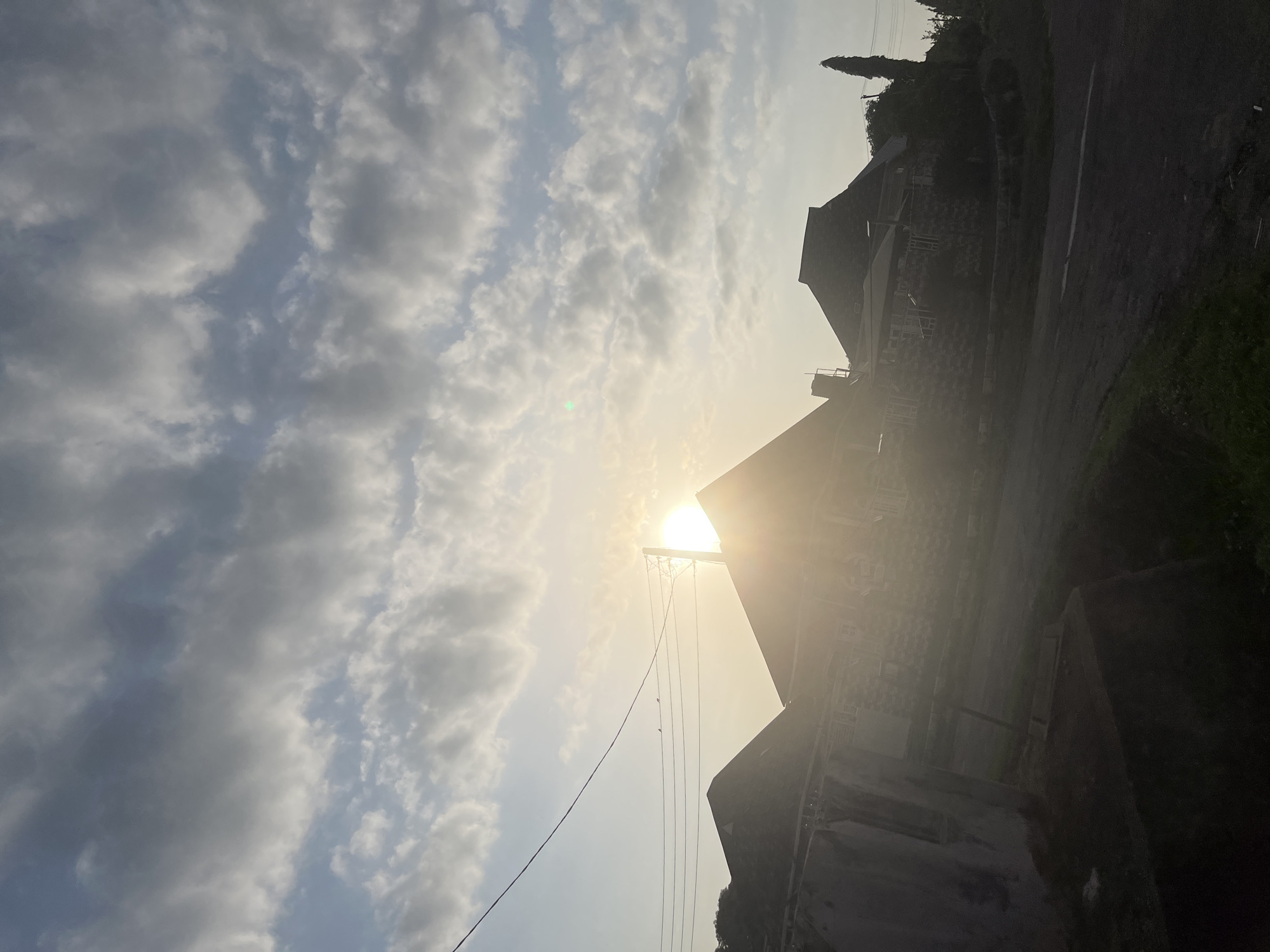 The image captures a street scene in Abuja, Nigeria, during sunrise or sunset. The sun is low on the horizon, casting a bright glow and lens flare across the sky. The sky is filled with a mix of scattered clouds, some appearing darker and heavier while others are lighter and more wispy. Silhouetted against the bright sky are the angular rooftops of residential buildings. Power lines and a utility pole are visible, with two small birds perched on one of the wires. The foreground shows a road with patches of grass and what appears to be a low wall or embankment on the left. The overall lighting suggests a hazy, diffused natural light typical of dawn or dusk.
