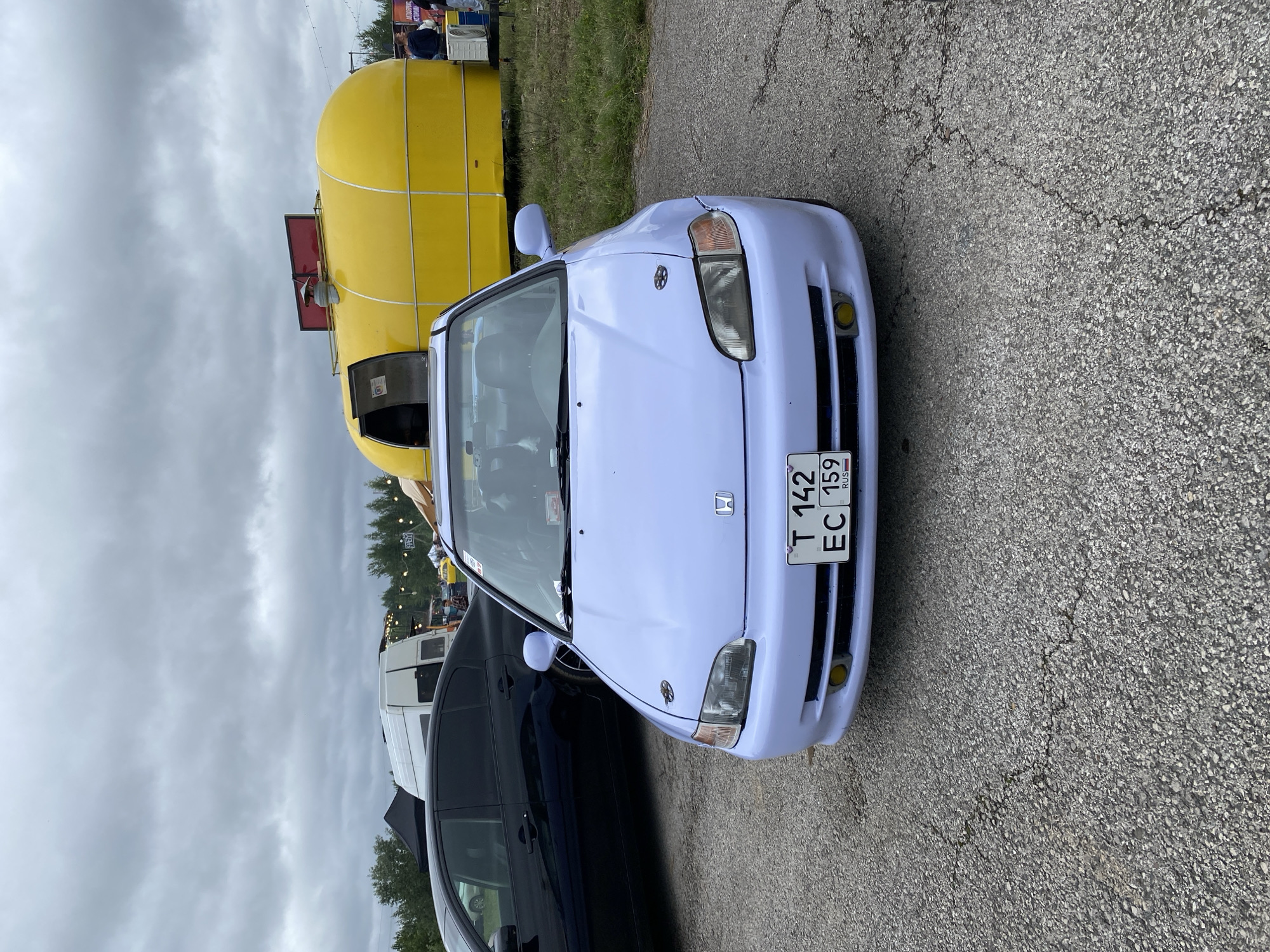 In Perm, Russia, under a cloudy sky, a light purple Honda Civic is parked on a cracked asphalt surface. The car, positioned at a slight angle facing the camera, features aftermarket modifications such as hood pins. Its license plate reads "T 142 EC 159 RUS". Behind the car, a black sedan is visible, and further in the background, a yellow, rounded food truck with an open door is present, along with a white van, string lights, and a line of trees. Several people are sparsely distributed in the background, near the food truck and van. The overall scene suggests an outdoor event or gathering in a semi-urban or industrial area.