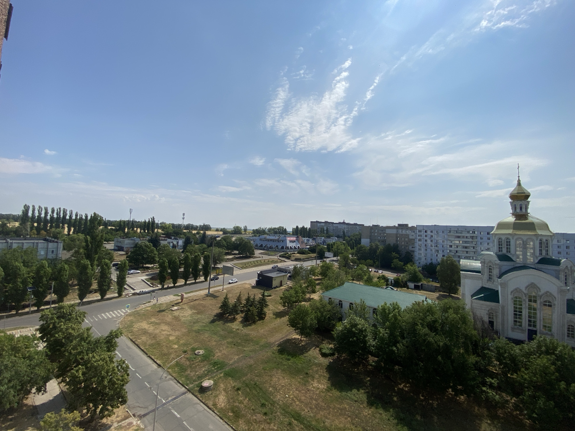 This is an aerial view of a city, identified as Pivdennoukrainsk, Ukraine. The scene depicts a sunny day with a bright blue sky and scattered white clouds. In the foreground, a paved road with lane markings curves through a grassy area dotted with trees. Several buildings are visible in the midground, including a large, white structure with a green roof and a prominent golden dome, which appears to be a church or religious building. Adjacent to it is another building with a green roof. Further in the background, apartment buildings and other structures stretch across the cityscape, interspersed with trees. A few vehicles are present on the roads, and a crosswalk is marked on one of the streets. The overall impression is of a residential and civic area on a clear day.