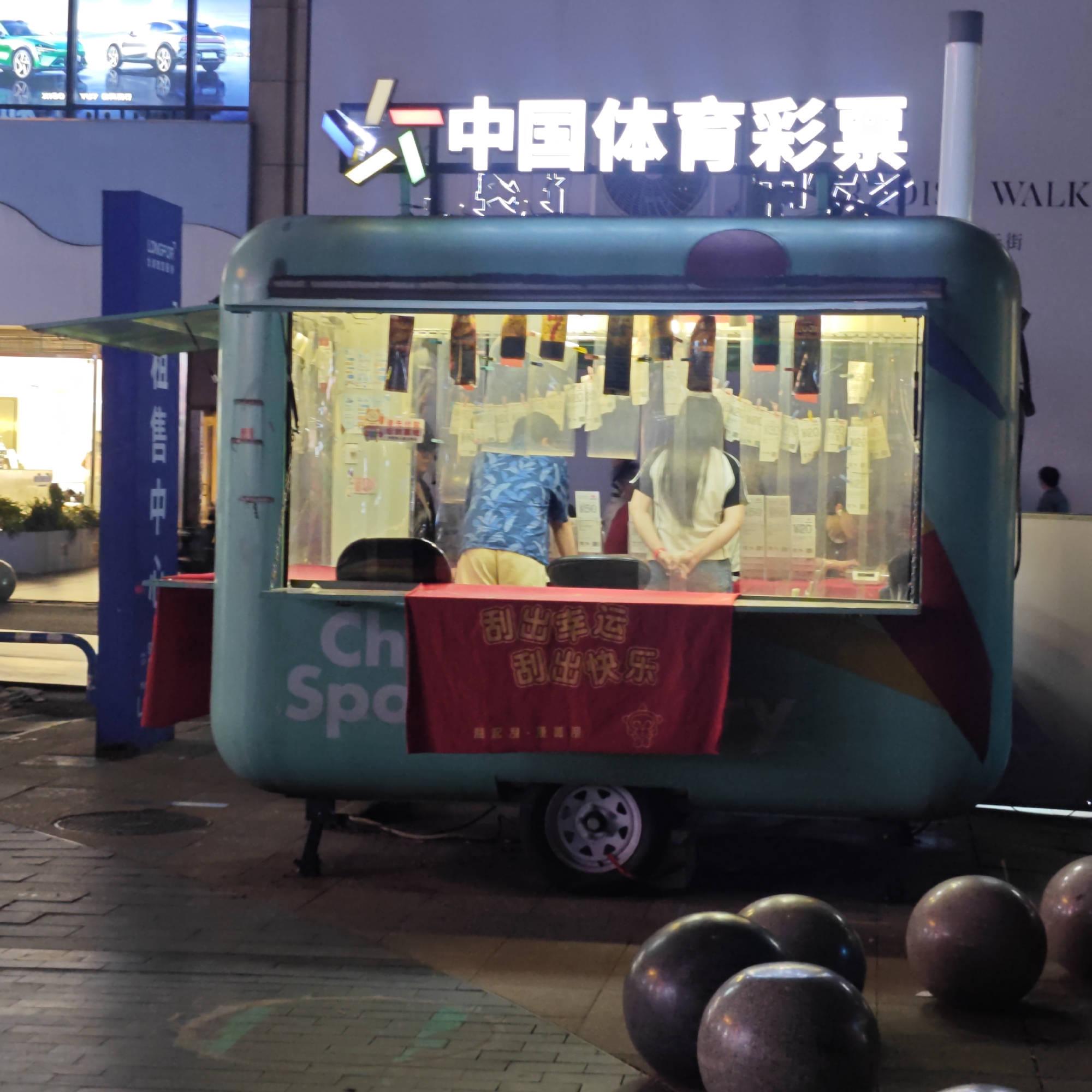 Here is a detailed description of the image:
**Overview:**
The image captures a scene at night, likely in a public area or commercial district in the city of Yuzhong District, China. The focal point is a small, mobile kiosk used for selling China Sports Lottery tickets.
**Details:**
* **Kiosk:** The kiosk is a blue-green color, and is located on wheels, indicating that it is mobile. It has a service window where customers can purchase lottery tickets. There is writing on the side of the kiosk, but part of it is obstructed. At the front of the kiosk is a red banner with Chinese characters on it; the characters are translated as "Scratch out lucky" and "Scratch out happy."
* **Lottery Tickets:** Inside the kiosk, lottery tickets can be seen hanging and displayed.
* **People:** Two people are visible inside the kiosk. A person with a blue shirt is to the left. To the right is a person with long hair, wearing a light-colored shirt.
* **Signage:** Above the kiosk, there is a neon sign that reads "中国体育彩票" (China Sports Lottery).
* **Environment:** The kiosk is set up in what appears to be a paved area. Decorative spheres or concrete balls are in the foreground.
* **Background:** In the background, there is a building with other businesses and signs, including "LONGFOR."
The image conveys a snapshot of a typical lottery retail operation in China.