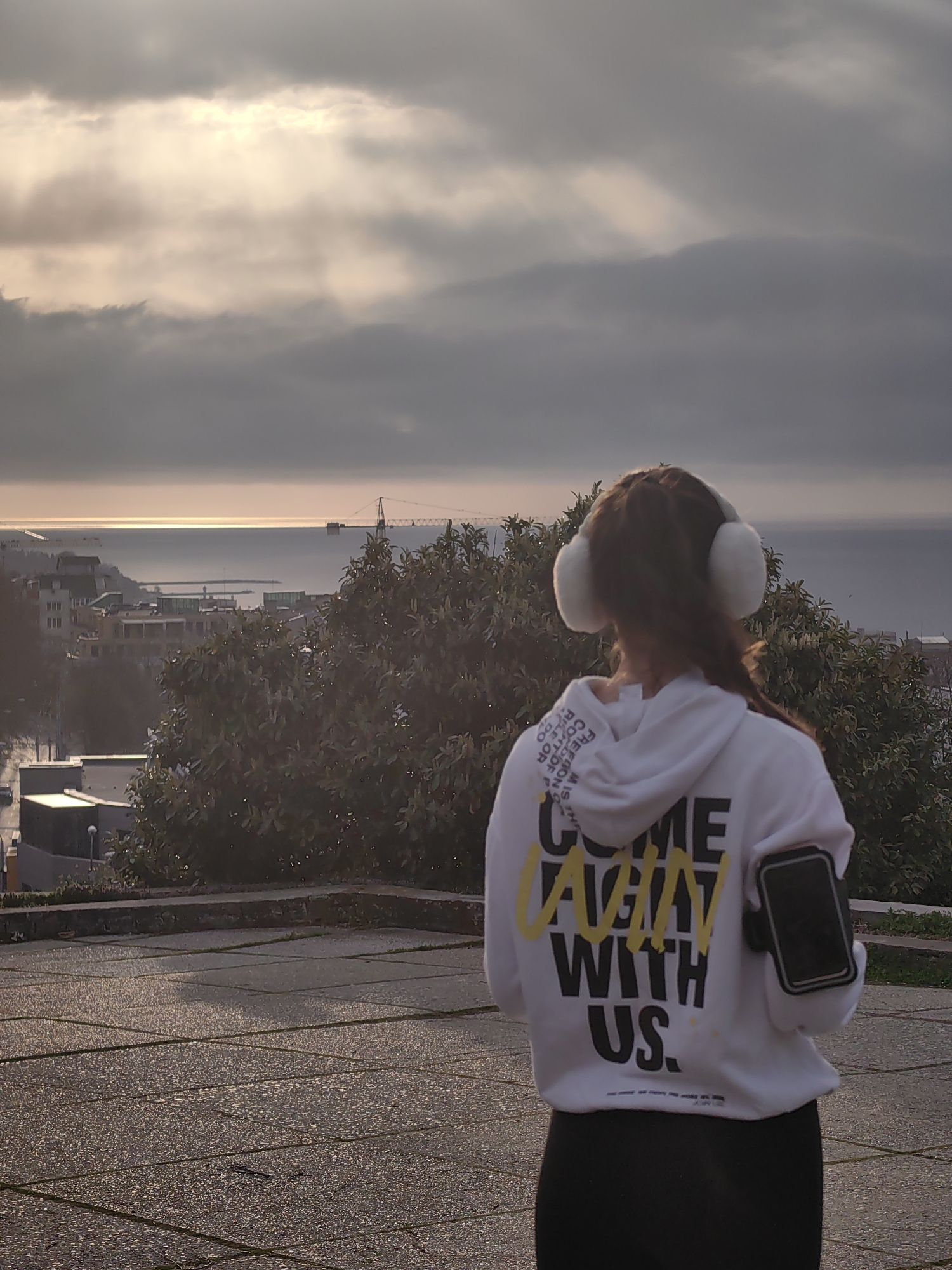 A woman stands on a paved area overlooking a city and the sea. She wears a white hoodie with black lettering and yellow accents, black leggings, and fluffy white earmuffs. A phone is strapped to her upper arm. The city appears to be Varna, Bulgaria, based on the architectural style and location on the Black Sea. The sky is overcast, with a hint of sunlight breaking through the clouds. In the background, a crane is visible on the coast. The woman is looking out towards the sea.