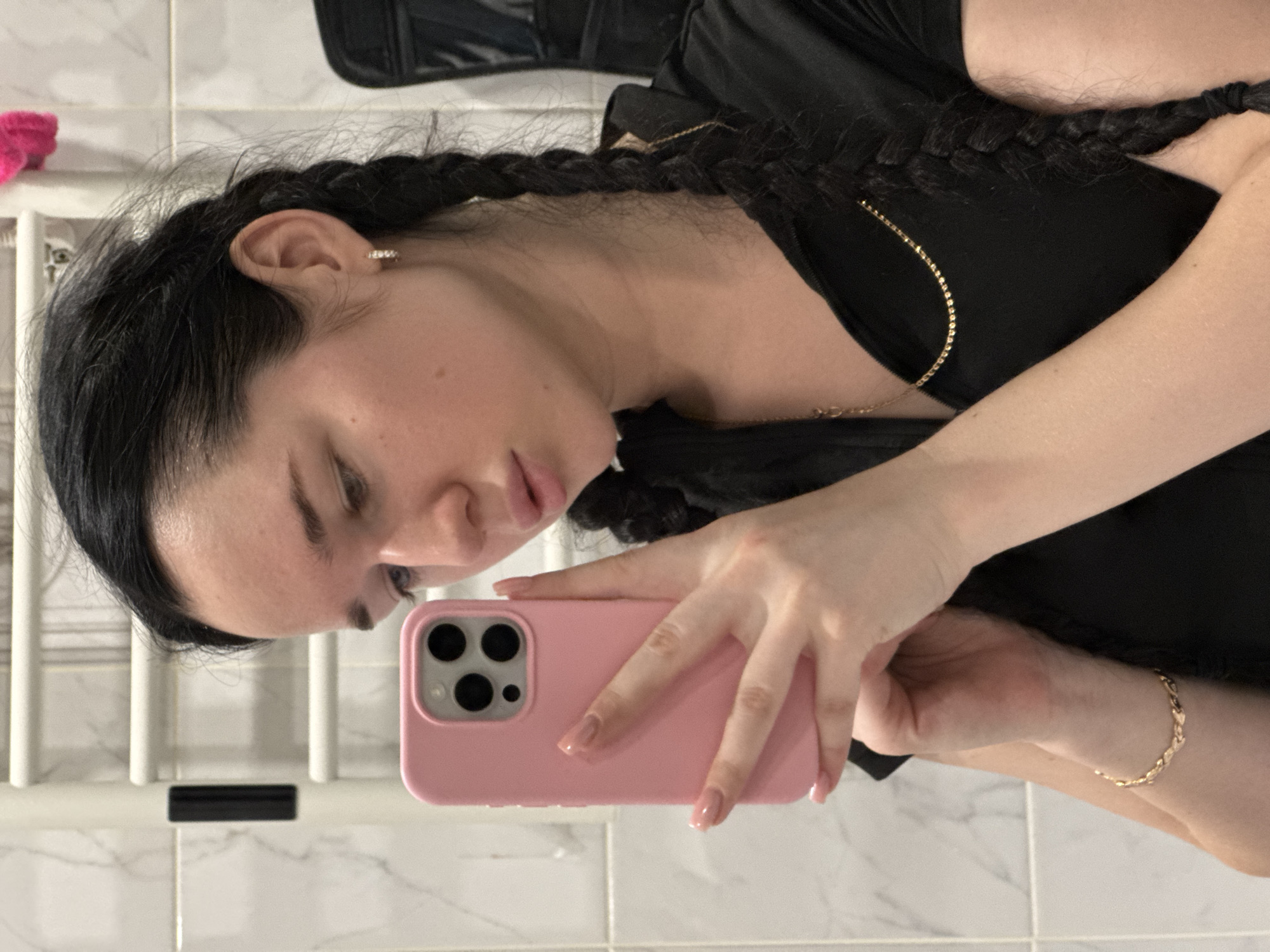 A young woman with dark hair in two braids takes a selfie in a bathroom. She is wearing a black top and a gold necklace. Her left hand holds a pink phone, and her right hand is visible, wearing a gold bracelet. The bathroom features white tiled walls and a silver towel rack. The image is a casual snapshot, likely taken in a residential setting.