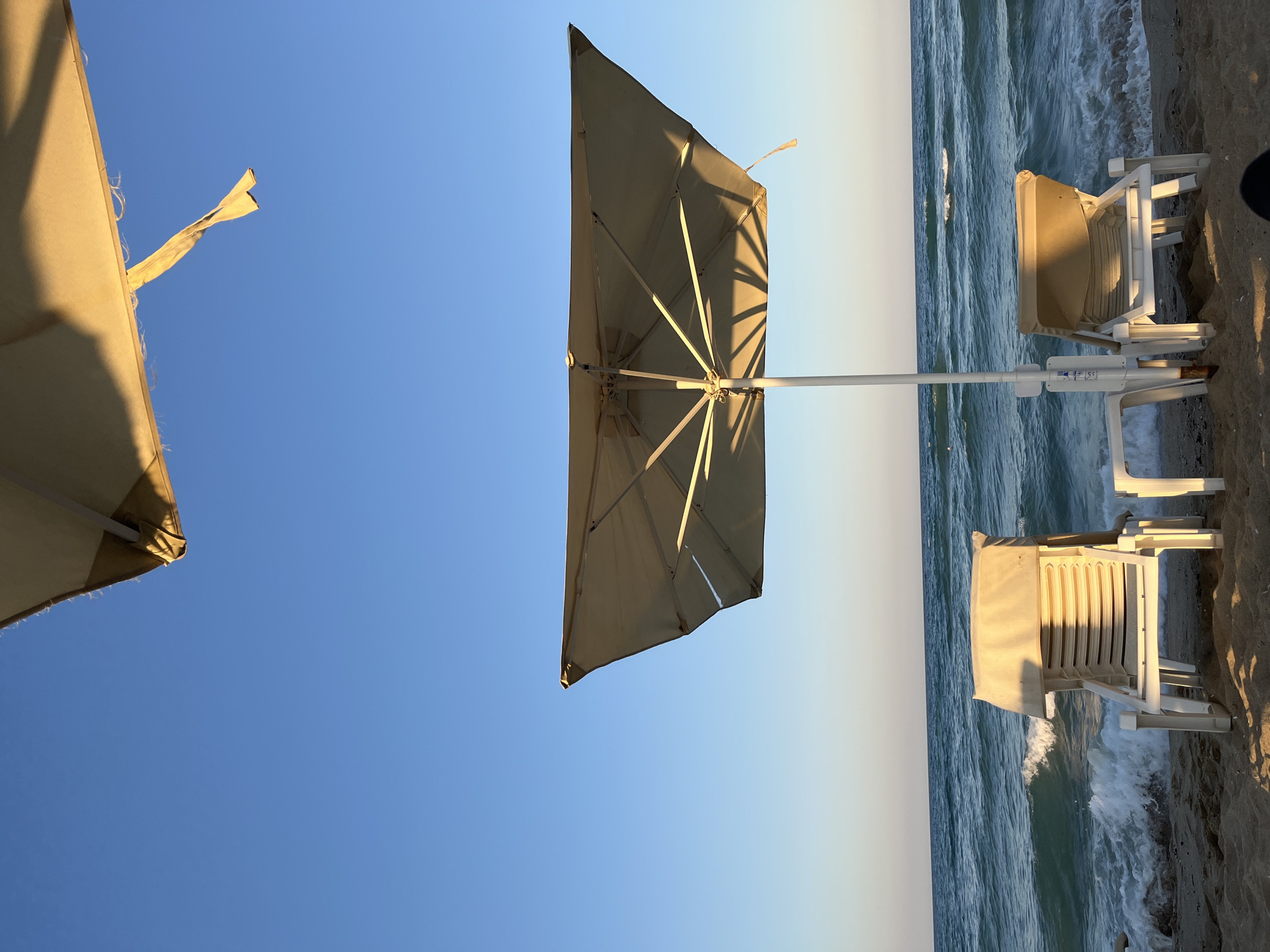Two white beach chairs and a large tan umbrella stand on a sandy beach facing the sea. The umbrella casts a shadow on the sand. The sea is a vibrant blue with white-capped waves rolling towards the shore. The sky is clear and blue. Part of another tan umbrella is visible at the top of the frame. The scene is located on the beach in Obzor, Bulgaria.