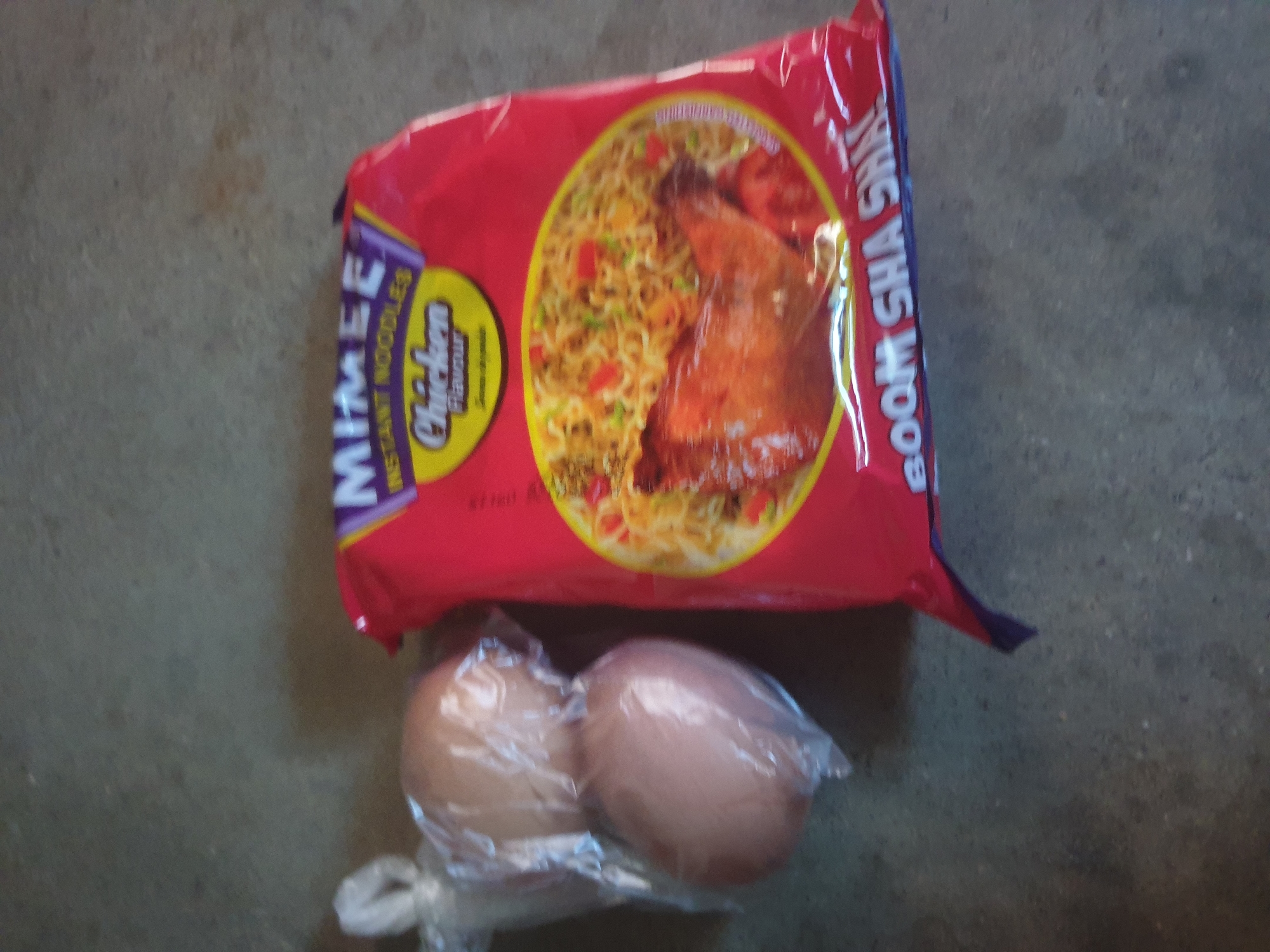 The image displays a red package of "MIMEE INSTANT NOODLES Chicken Flavour" and two light brown eggs contained within a clear plastic bag. Both items rest on a flat, textured, grayish-beige surface, resembling concrete, in an unknown location. The noodle package features a large circular graphic depicting cooked instant noodles with red and green garnishes, alongside a piece of cooked chicken. The eggs are situated to the left of the noodle package, slightly elevated in the frame. The plastic bag holding the eggs is loosely tied at the top. The surface exhibits minor variations in color and texture. The lighting is diffused and even, with no harsh shadows.