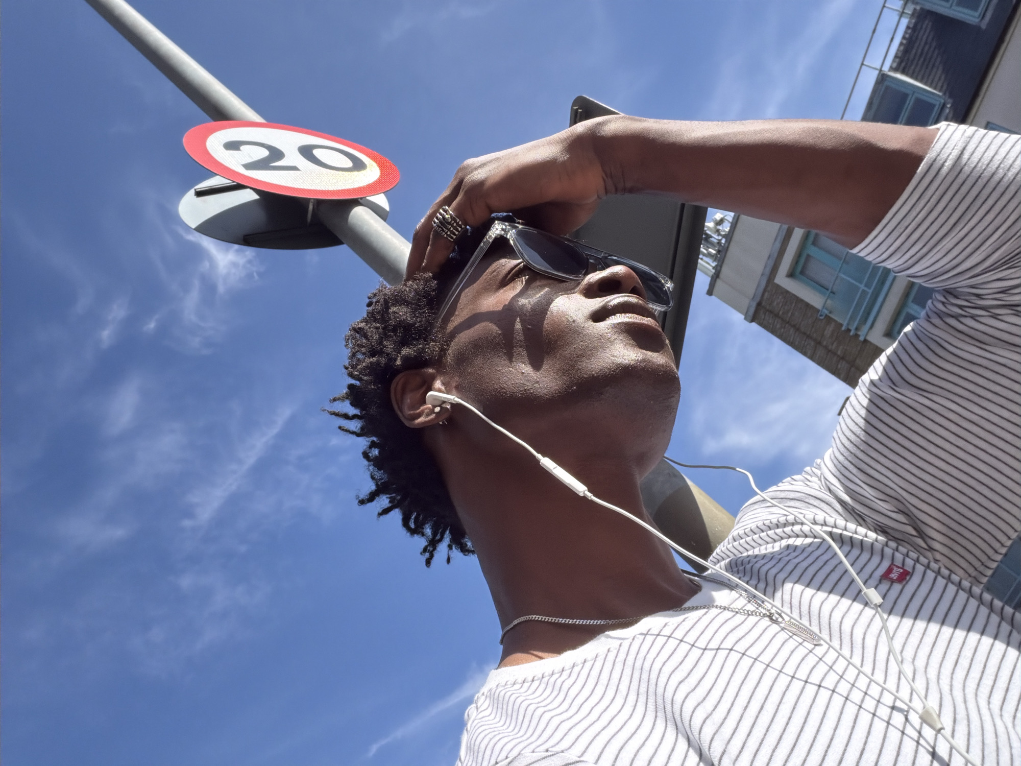 A low-angle shot reveals a dark-skinned individual wearing a white shirt with black horizontal stripes, a silver necklace, and white earphones. Sunglasses are perched on the individual's face, held in place by their hand. Above, a road sign indicating a speed limit of 20 is visible, mounted on a gray pole against a bright blue sky with scattered clouds. In the background, parts of a building with blue-framed windows can be seen. The location is possibly within the city of London, United Kingdom.