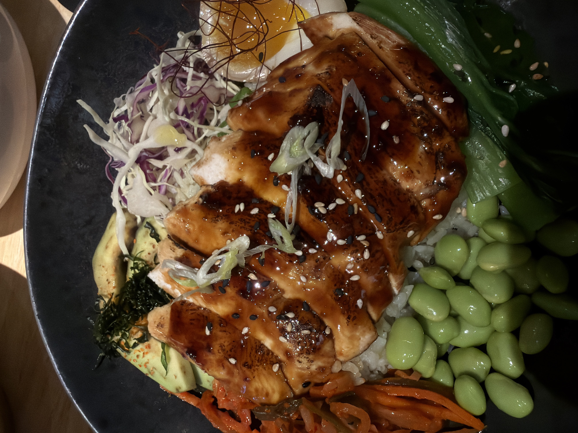 A close-up shot depicts a portion of a dark-colored bowl filled with an assortment of food items, including sliced teriyaki chicken, shredded white and purple cabbage, sliced avocado, cooked edamame, kimchi, a soft-boiled egg, and stir-fried bok choy. The meal is likely served in North Kuta, Indonesia.