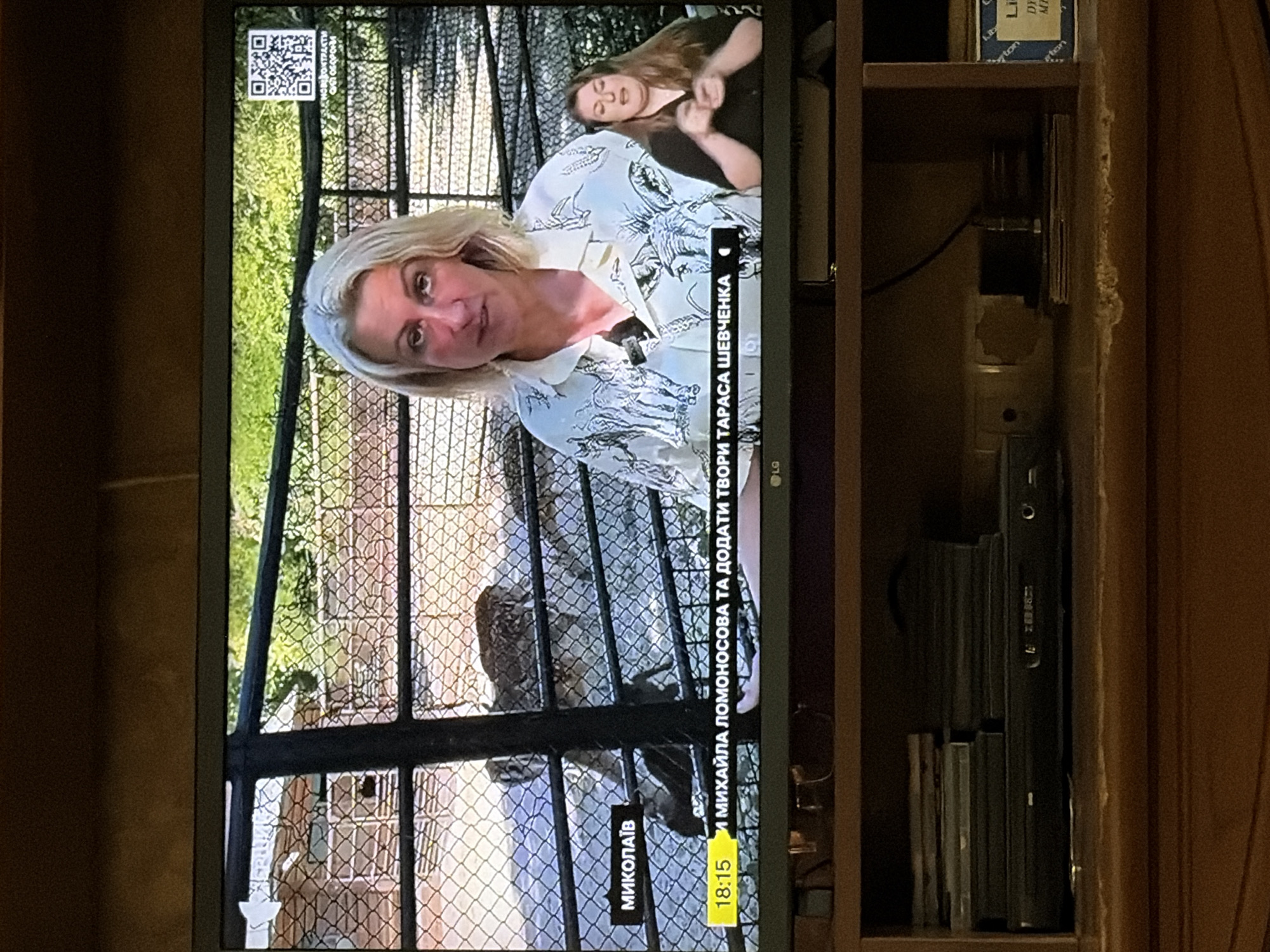 The image shows a television screen displaying a news broadcast. The broadcast features a woman speaking directly to the camera, with a smaller window showing a sign language interpreter on the right. The background of the main broadcast appears to be a zoo or animal enclosure, with fencing and greenery visible. In the bottom left corner of the screen, there is a yellow banner with black text that reads "МИКОЛАЇВ" and "18:15" followed by Ukrainian text that likely indicates the news segment's topic. In the lower part of the image, below the television, a wooden cabinet is visible, with some electronic devices and stacked items inside. The overall scene appears to be a living room or common area where the television is being watched.