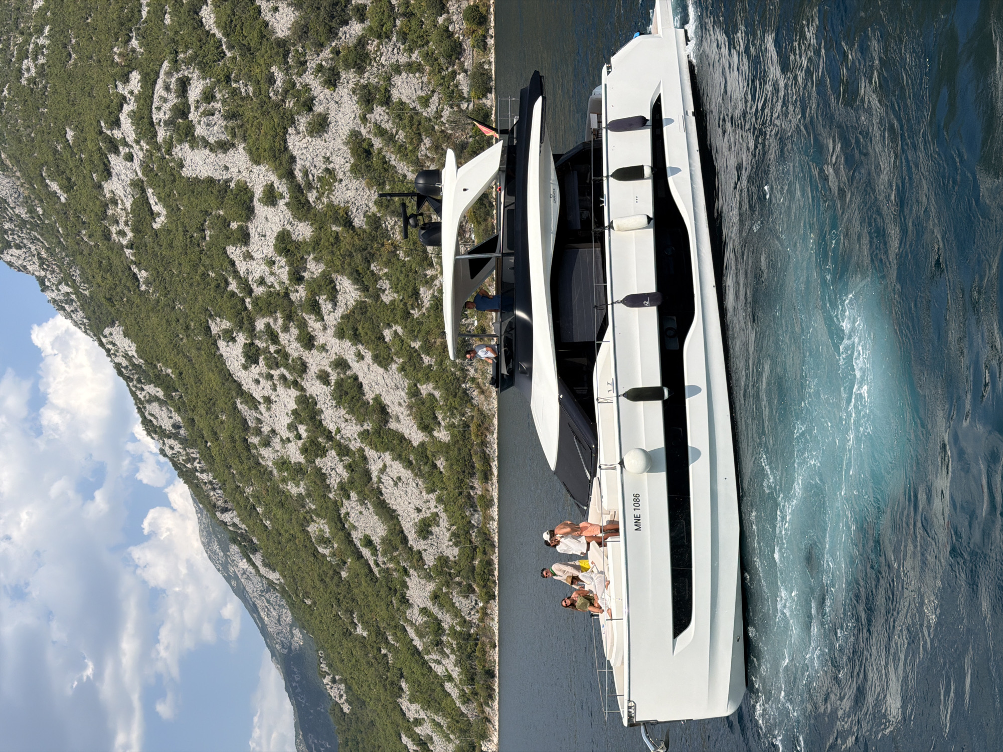A wide, low-angle shot captures a modern, white yacht cruising on a deep blue body of water, with a lush, green mountain range providing a dramatic backdrop under a partly cloudy sky. The yacht, identified by the registration "MNE 1086," has a sleek design with black tinted windows and several people visible on its deck. Four individuals are standing near the bow, appearing to converse and observe the scenery, while two others are situated further back on the upper deck. The water surrounding the yacht is churned up, creating white foam and swirls, indicating movement. The mountains are densely covered in vegetation, with rocky outcrops visible, suggesting a rugged and natural environment. The scene conveys a sense of leisure and luxury against a stunning natural landscape, likely within the Bay of Kotor, near the historic town of Perast, Montenegro.