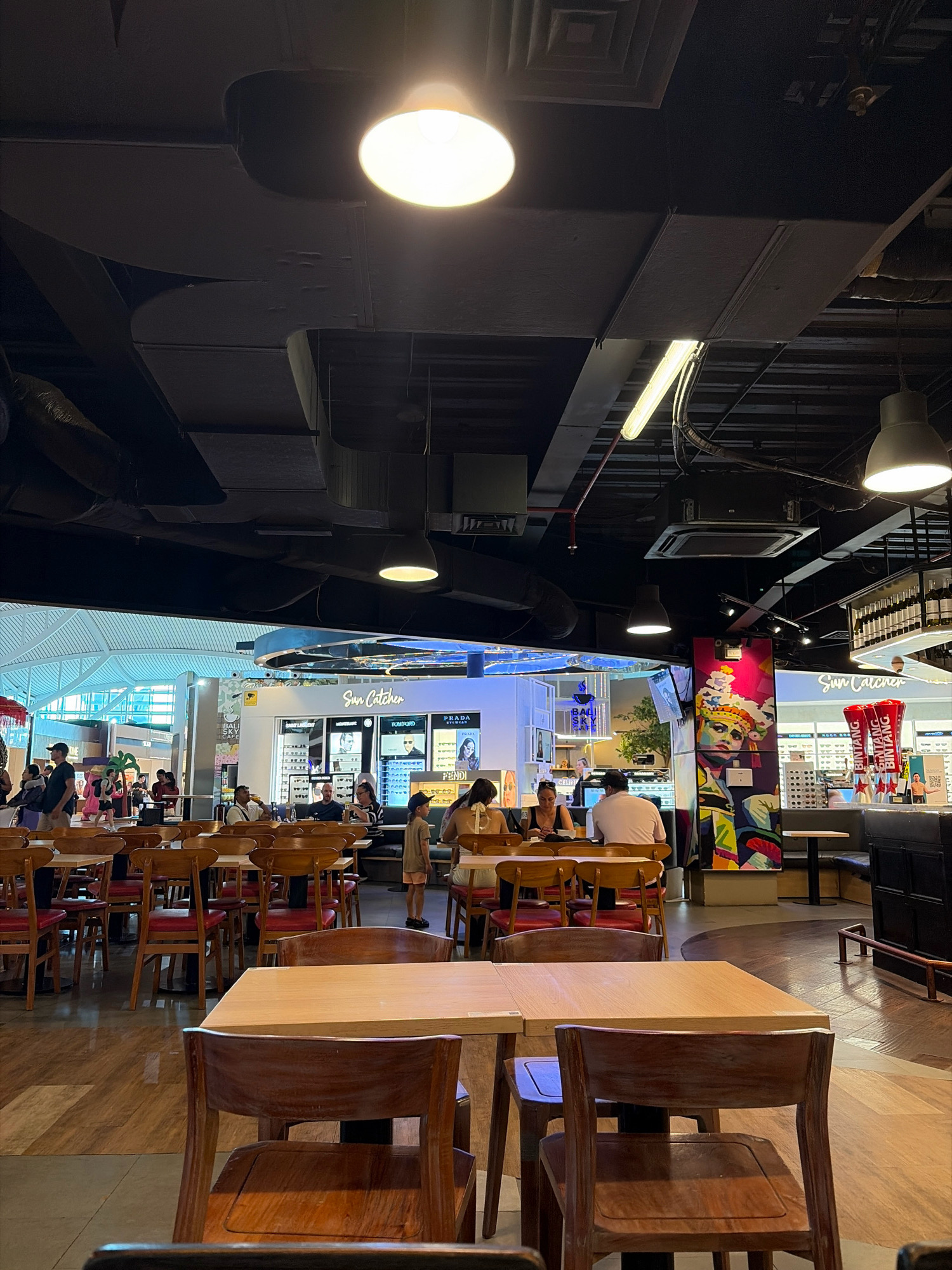 An indoor scene, possibly an airport or a large shopping mall in Kuta, Indonesia, is captured from a slightly elevated perspective, looking towards a bright, open area with large glass windows. In the foreground, several wooden tables with red cushioned chairs are set up, suggesting a dining or waiting area. Some of the tables are empty, while others have customers seated, engaged in conversations or looking at their phones.
In the midground, a brightly lit store with various branded sunglasses displays, including "Tom Ford" and "Prada," is visible. Adjacent to this, a colorful mural featuring a woman's face adorns a pillar. Beyond this area, a spacious, light-filled atrium with a modern architectural design is visible through the large glass windows, with people walking around. The ceiling is a dark, industrial design with exposed ductwork and multiple light fixtures, some of which are brightly illuminated. The floor is a combination of polished wood and reflective surfaces. The overall atmosphere appears to be bustling and commercial.