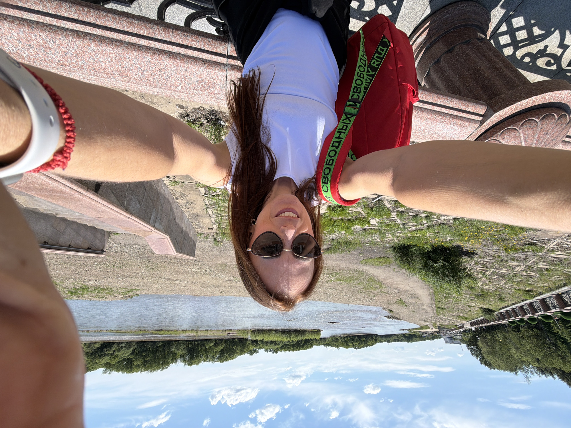A woman with long, reddish-brown hair and sunglasses takes a selfie while standing on a promenade overlooking a river on a sunny day. She is wearing a white sleeveless top and a red backpack with a green strap that says "CBOROWHH". To her left, there is a decorative stone railing and a stone pillar. In the background, a river flows, with a tree-lined bank on the opposite side. The promenade has a paved walkway with some patches of grass and yellow flowers. The sky is blue with scattered white clouds. The location is the Amur River embankment in Birobidzhan, Russia.