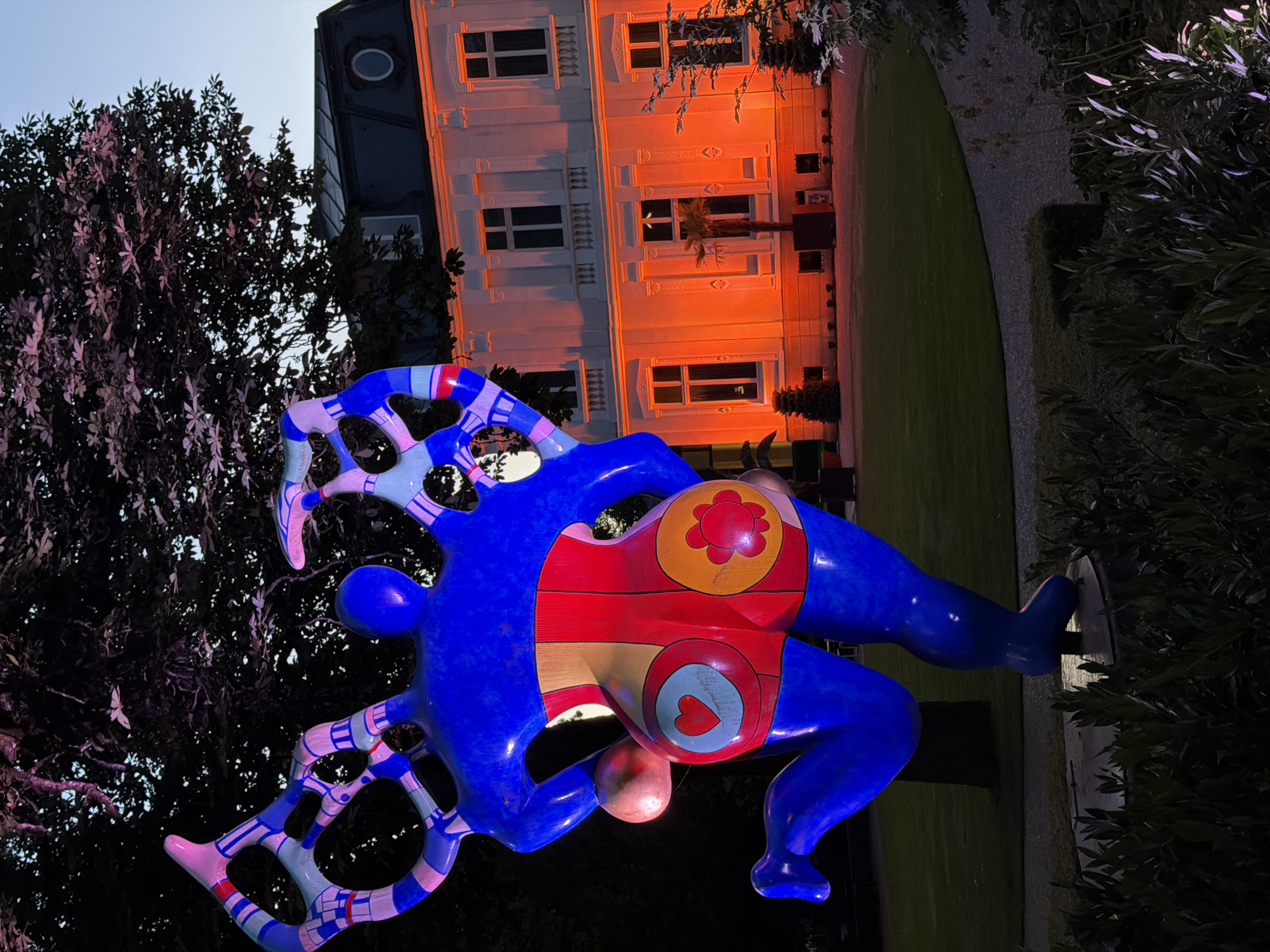 An illuminated, large, blue, stylized female sculpture with red and yellow patterns on its lower body is situated in a garden at dusk. The sculpture is in a dynamic pose, with one leg bent and the other extended. The sculpture's upper limbs are abstract and wing-like, with blue and pink geometric patterns. In the background, a large, white building with a mansard roof is illuminated with orange light. The building has multiple windows and decorative architectural elements. Trees and manicured bushes are visible around the sculpture and the building. The scene appears to be outdoors, possibly in a park or a public garden. The location is the Villa Vauban - Musée d'Art de la Ville de Luxembourg in Luxembourg City, Luxembourg.