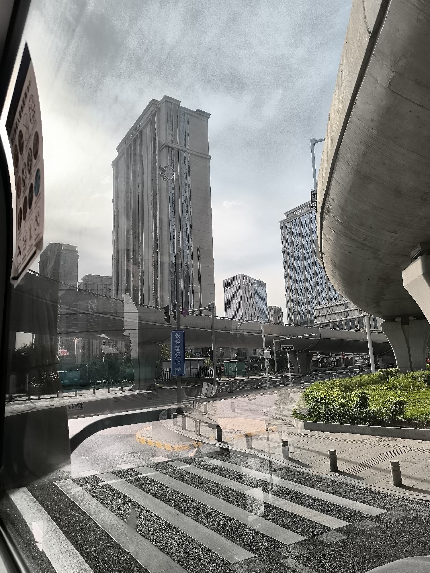 The image depicts an urban scene in Ürümqi, China, viewed from inside a vehicle, likely a bus, as indicated by the reflections on the glass. The foreground features a wet asphalt road with a pedestrian crossing marked by white stripes. To the right, a sidewalk with bollards is visible, bordered by a grassy area.
In the midground, a multi-lane road curves, and an elevated highway structure with concrete supports dominates the right side of the frame. Traffic lights and street signs are present. A blue sign with white Chinese characters, possibly indicating a bus station, is positioned near the traffic lights. In the background, several modern high-rise buildings rise against a cloudy sky. The overall atmosphere is overcast, suggesting a potentially rainy or humid day. Reflections on the vehicle's windows show parts of the interior, including a poster or informational leaflet with circular graphics. The image captures a typical urban environment with infrastructure, buildings, and signs characteristic of a Chinese city.