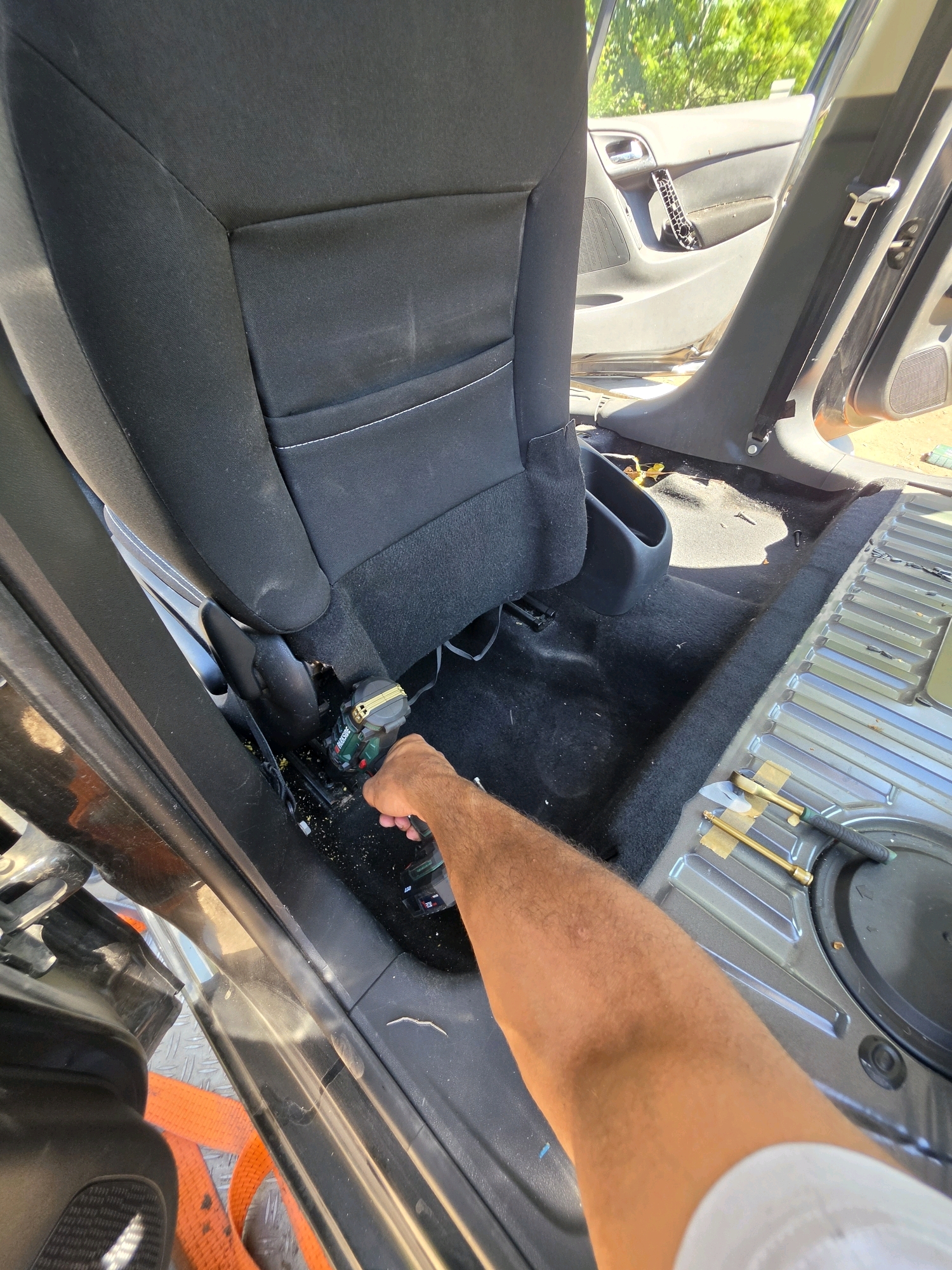 The image depicts the interior of a vehicle, specifically the rear passenger area, in the city of Varna, Bulgaria. A person's arm and hand are visible on the right side of the frame, holding a green and black power tool, likely a drill, which is being used near the base of a black fabric car seat. The seat is partially obscured, but its general shape and material are discernible. The floor of the car is covered in black carpet, with some debris and dust scattered around the area where the tool is being operated. To the right of the carpeted floor, a metallic ribbed surface is visible, indicating the undercarriage or a structural component of the vehicle. In the background, the open door of the car reveals a glimpse of the exterior, with green foliage and sunlight suggesting an outdoor environment. The overall impression is that of a car interior undergoing some form of maintenance or repair.