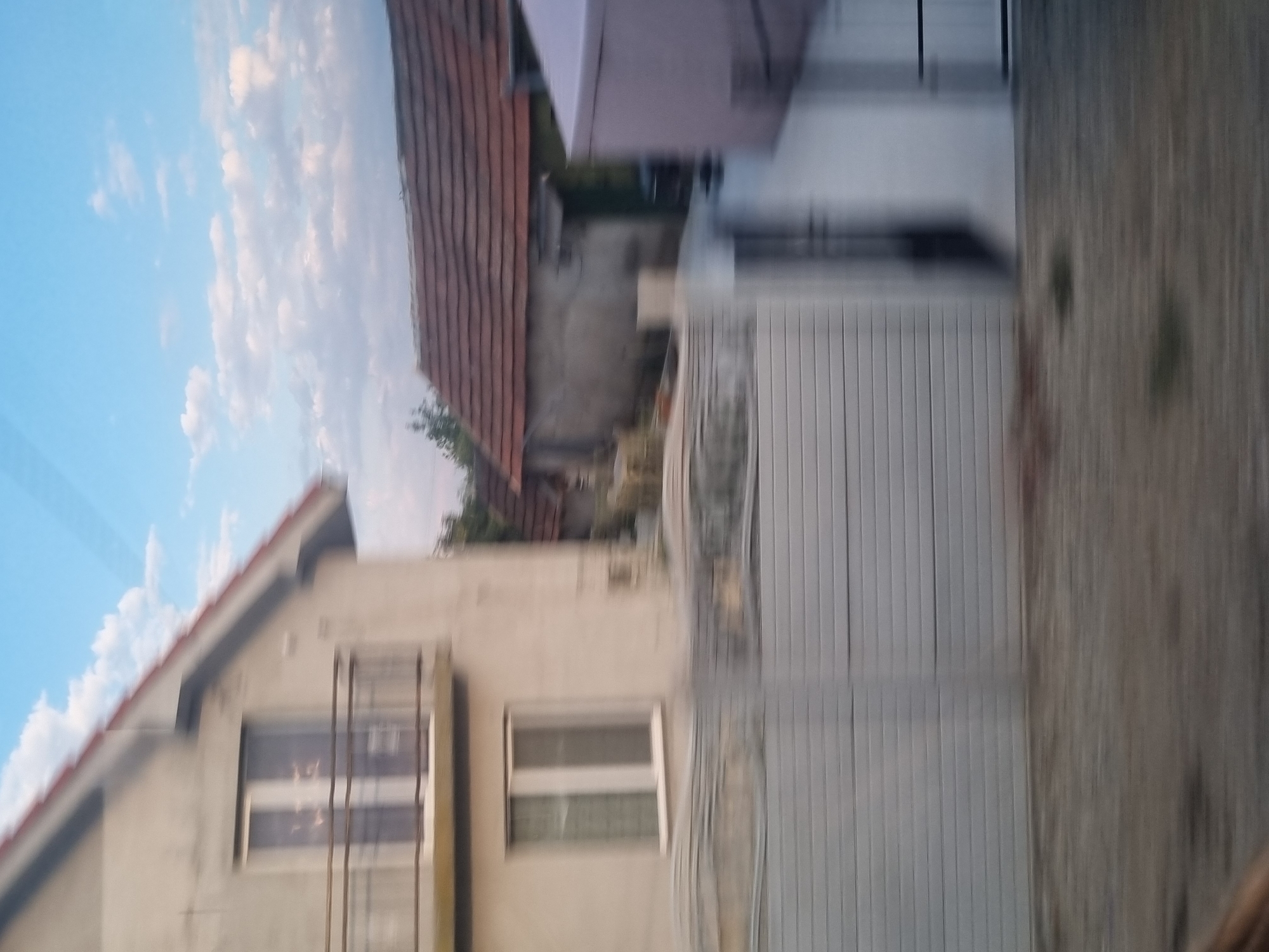 The image captures a blurred view of a residential area, likely in Inand, Romania, with a focus on several houses and a fence. The foreground consists of a gray, horizontally slatted fence. Behind the fence, a pale-colored house with a balcony and two windows is visible on the left. To the right, a house with a red-tiled roof and a concrete wall is depicted. Further to the right, a light purple structure, possibly a shed or outbuilding, is present. The sky above is blue with scattered white clouds. The motion blur suggests the image was taken from a moving vehicle.