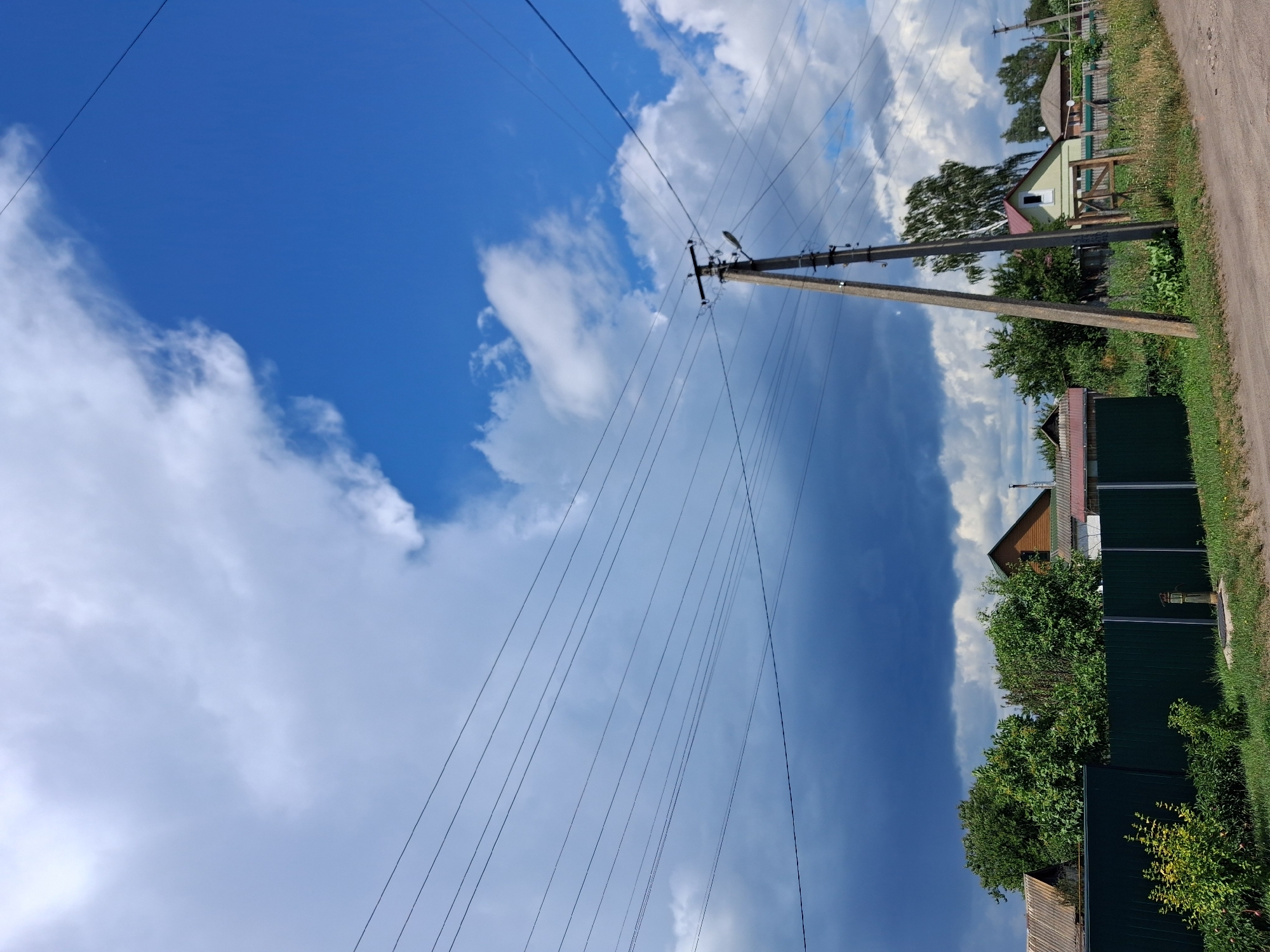 The image depicts a rural landscape with a backdrop of a dramatic sky. Overhead, numerous power lines crisscross the frame, strung between a tall concrete utility pole on the right and an unseen point to the left. The sky is a mix of bright blue and dark, ominous clouds, suggesting an approaching storm or recent rain.
In the foreground, a dark green metal fence runs horizontally across the image, separating the road from a residential area. Beyond the fence, several houses and trees are visible. The houses appear to be modest, with pitched roofs. Dense green foliage from trees and bushes fills the space between the houses and obscures parts of the structures. The ground in front of the fence is covered in green grass, with a dirt road running along the bottom edge of the frame. A yellow, metallic object, possibly a well or a pump, is visible near the fence line.
The scene is devoid of any people or animals. The overall impression is one of a quiet, semi-developed rural area under a dynamic sky. The location is identified as Krolevets, Ukraine, suggesting a setting in that specific region.