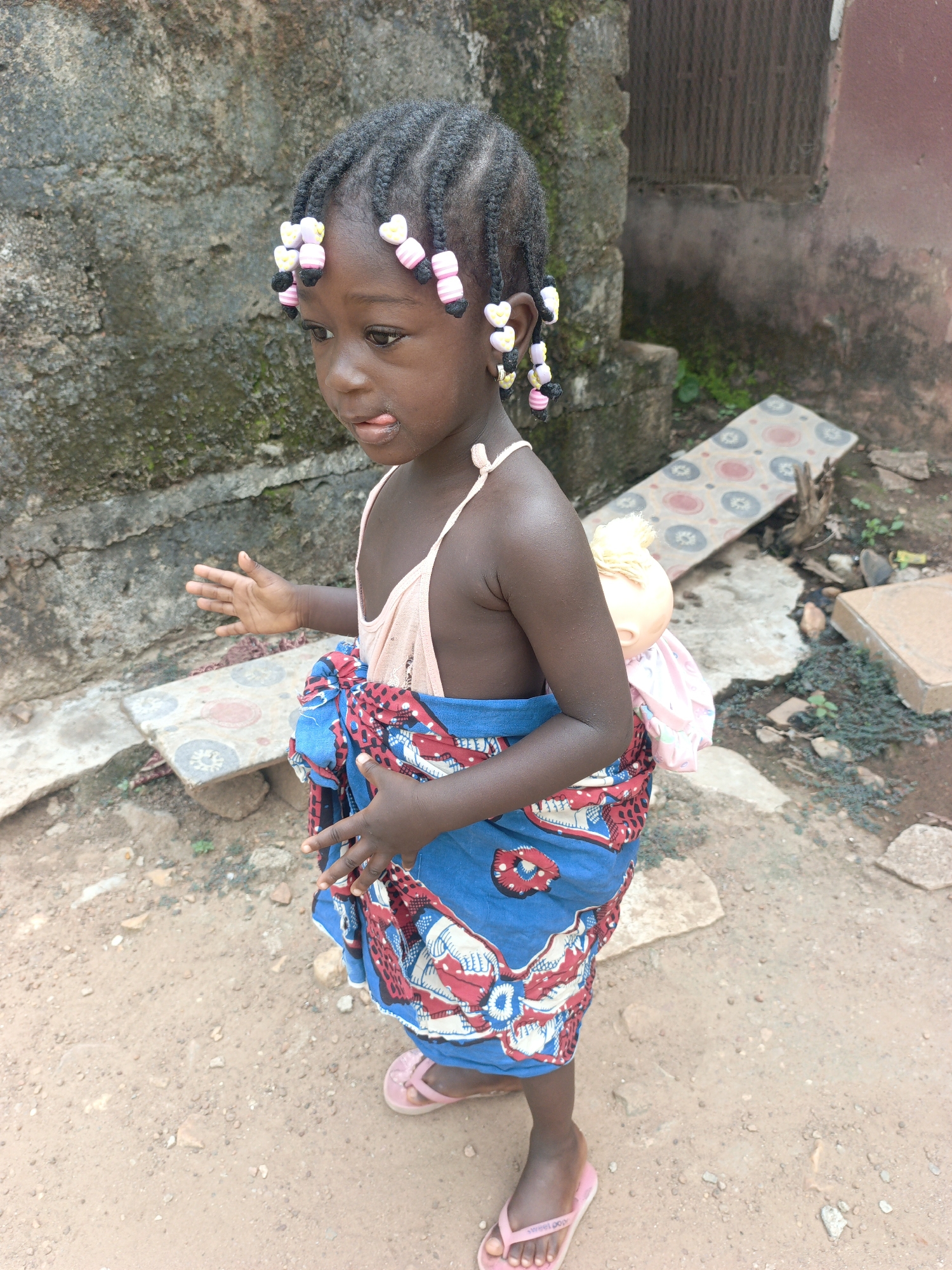 A young girl stands outdoors on a dirt surface. She has dark skin, dark hair braided with colorful beads, and is wearing a light pink tank top, a blue and red patterned wrap-around skirt, and pink sandals. A doll is secured on her back by the wrap. The background features a weathered concrete wall, a few small plants, and fragmented tiles. The image appears to be taken in Akwanga, Nigeria.