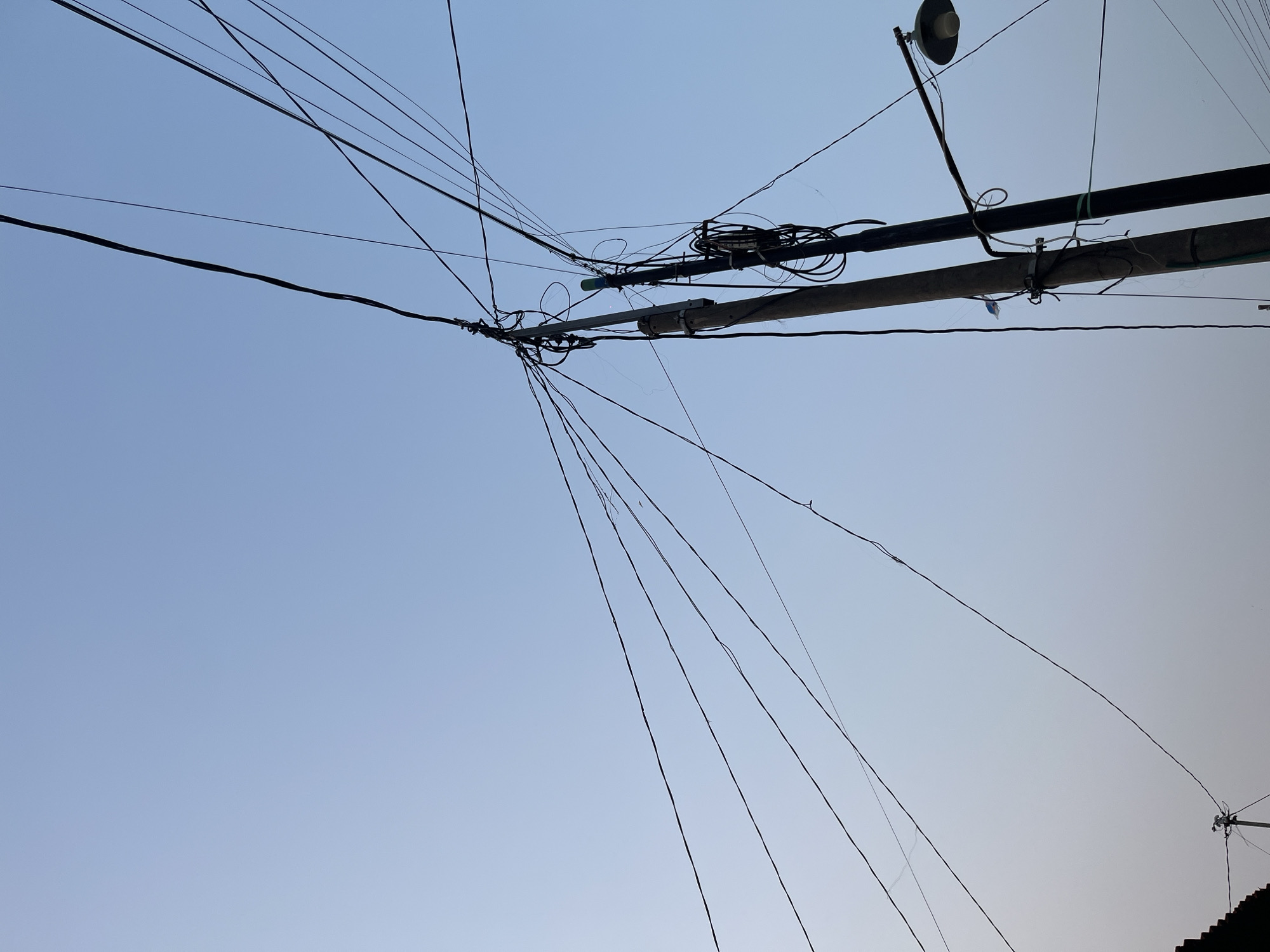 The image is a low-angle, full-frame shot of a telephone pole with numerous wires extending into a pale blue sky. The pole, made of concrete, is situated in Pekalongan, Indonesia. Attached to the pole is a street lamp. The wires are a tangled mess, indicative of urban infrastructure in Indonesia, which often features complex networks of overhead cables. The sky is clear and hazy, suggesting it might be early morning or late afternoon. There are no people visible in the frame, and the focus is solely on the utility pole and the dense web of electrical and telecommunication lines. The overall impression is one of a typical street scene in a densely populated area of Pekalongan.