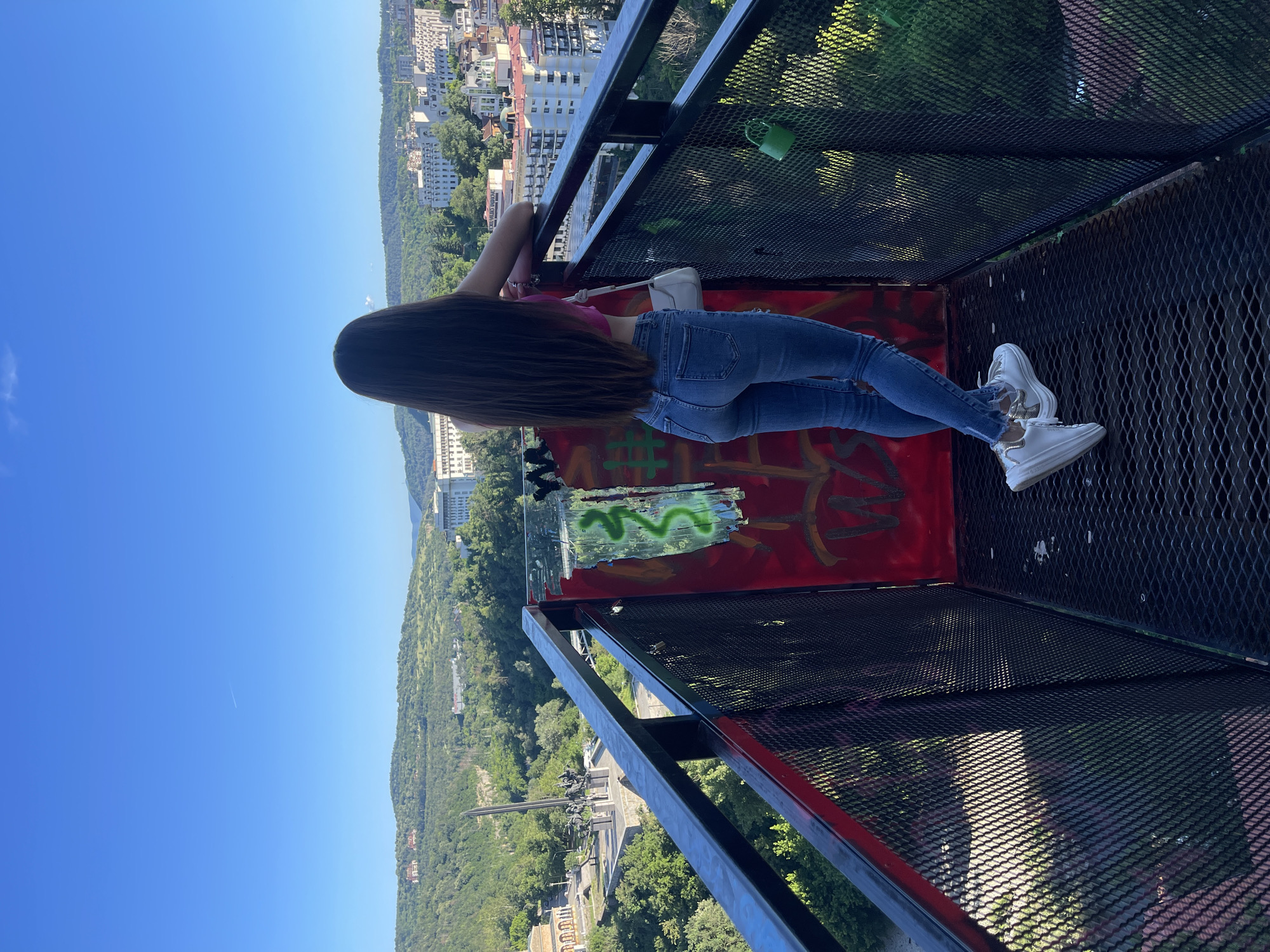 A young woman with long, dark hair stands on a metal platform, facing away from the camera, looking out at a city in the distance. She is wearing a pink crop top, blue jeans, and white sneakers. The platform has a black metal railing with red and green graffiti on a metal panel behind her. A white purse hangs from her left shoulder. In the distance, a city skyline is visible, with buildings nestled among rolling green hills under a clear blue sky. A monument with a tall spire is visible in the middle ground. A green padlock is attached to the railing. The scene is captured in daylight.